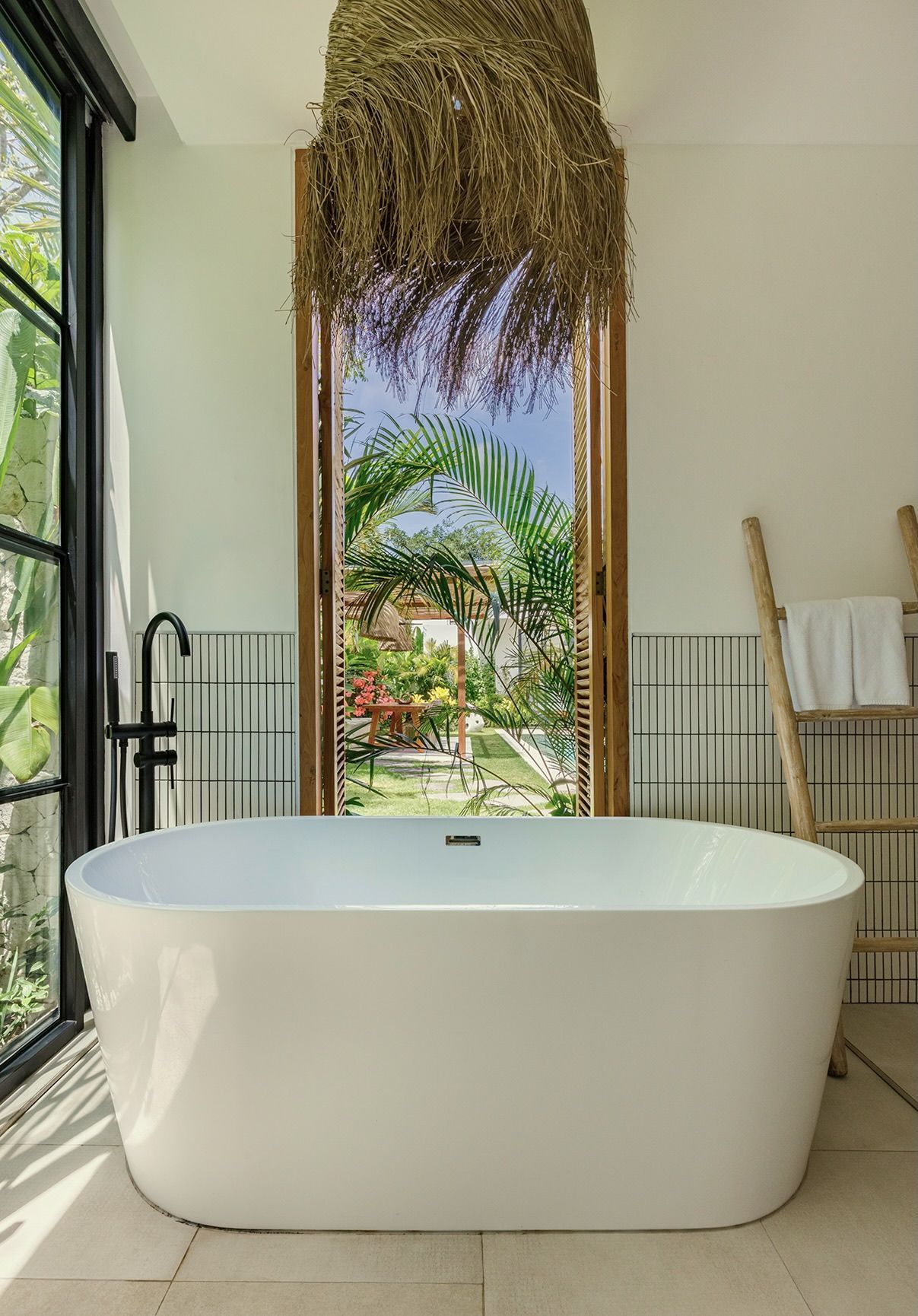 Freestanding white bathtub in a bright bathroom with open doors to a tropical garden