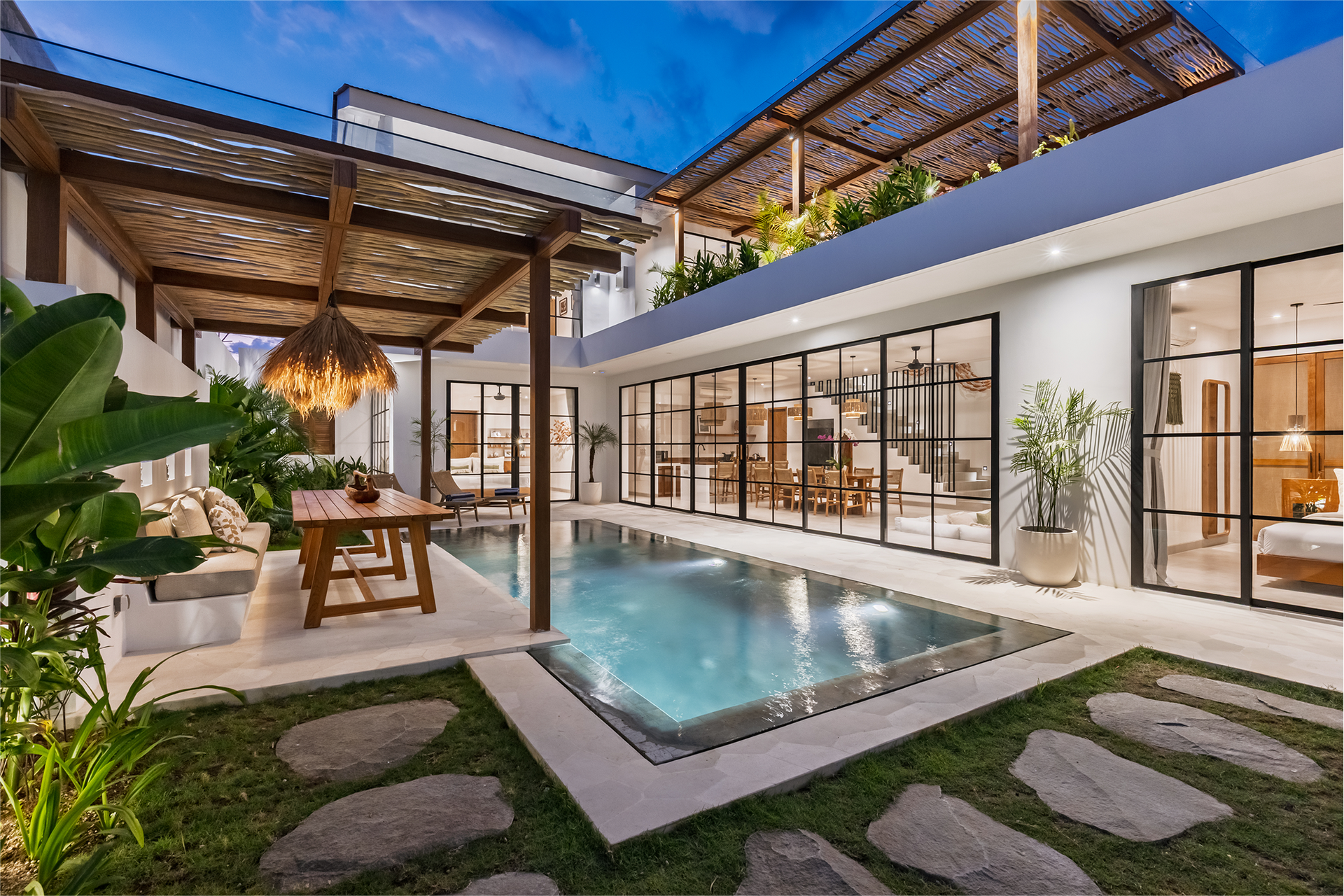 Modern patio with a small pool, glass-walled house, lounge area, and tropical plants at dusk