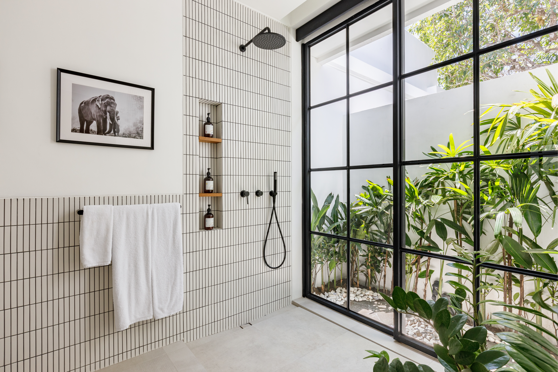 Modern tiled bathroom with a walk-in shower, black-framed glass door, towel, and greenery outside the window