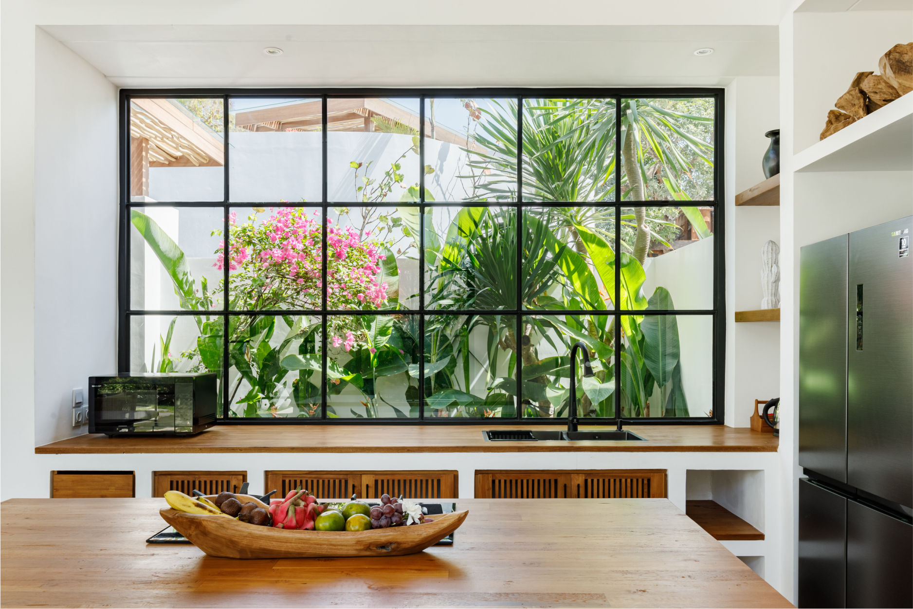 Bright kitchen with a wooden table, fruit bowl, and a large window overlooking lush greenery and pink flowers