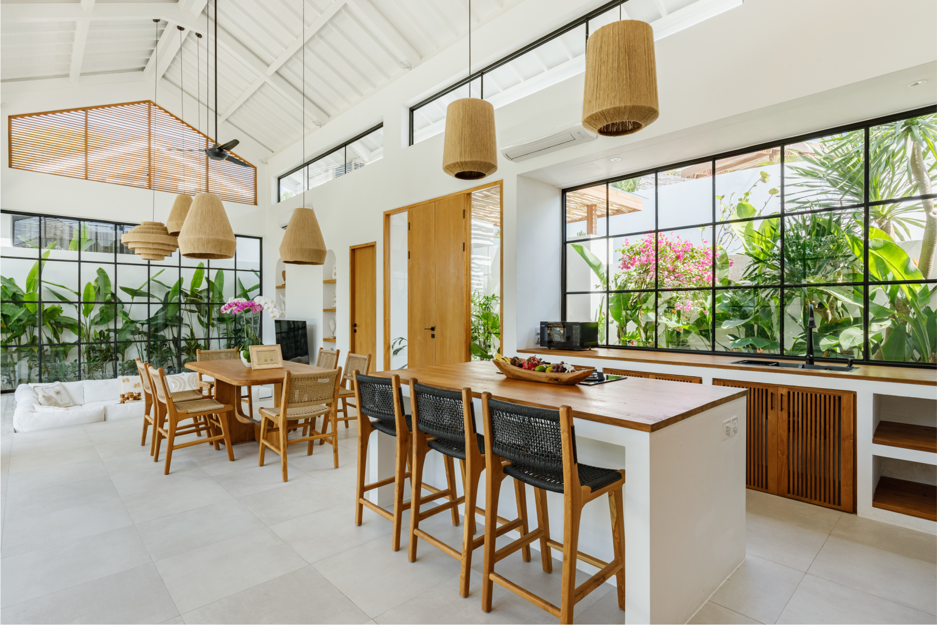 Bright tropical kitchen and dining area with white walls, wooden furniture, and large windows overlooking greenery
