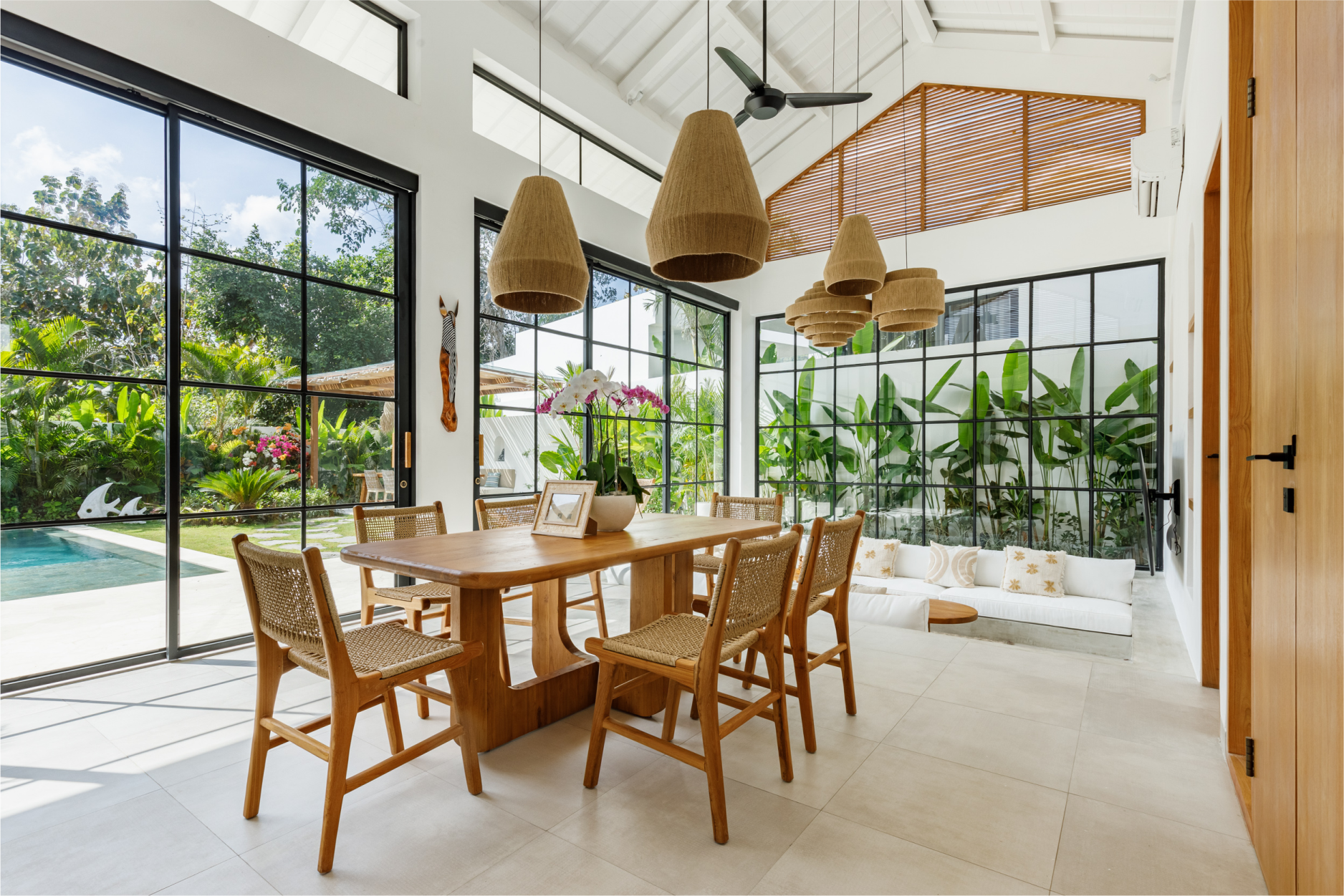 Bright dining room with wooden table, wicker chairs, and large windows overlooking a pool and lush garden