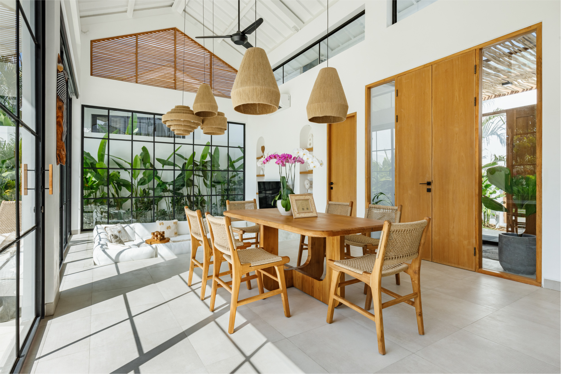 Bright dining room with wooden table, woven chairs, hanging lights, and large windows overlooking greenery