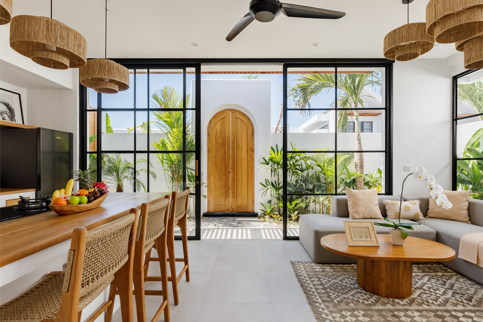 Bright, open living room with wicker dining table, large windows, wood door, and cozy beige sofa.