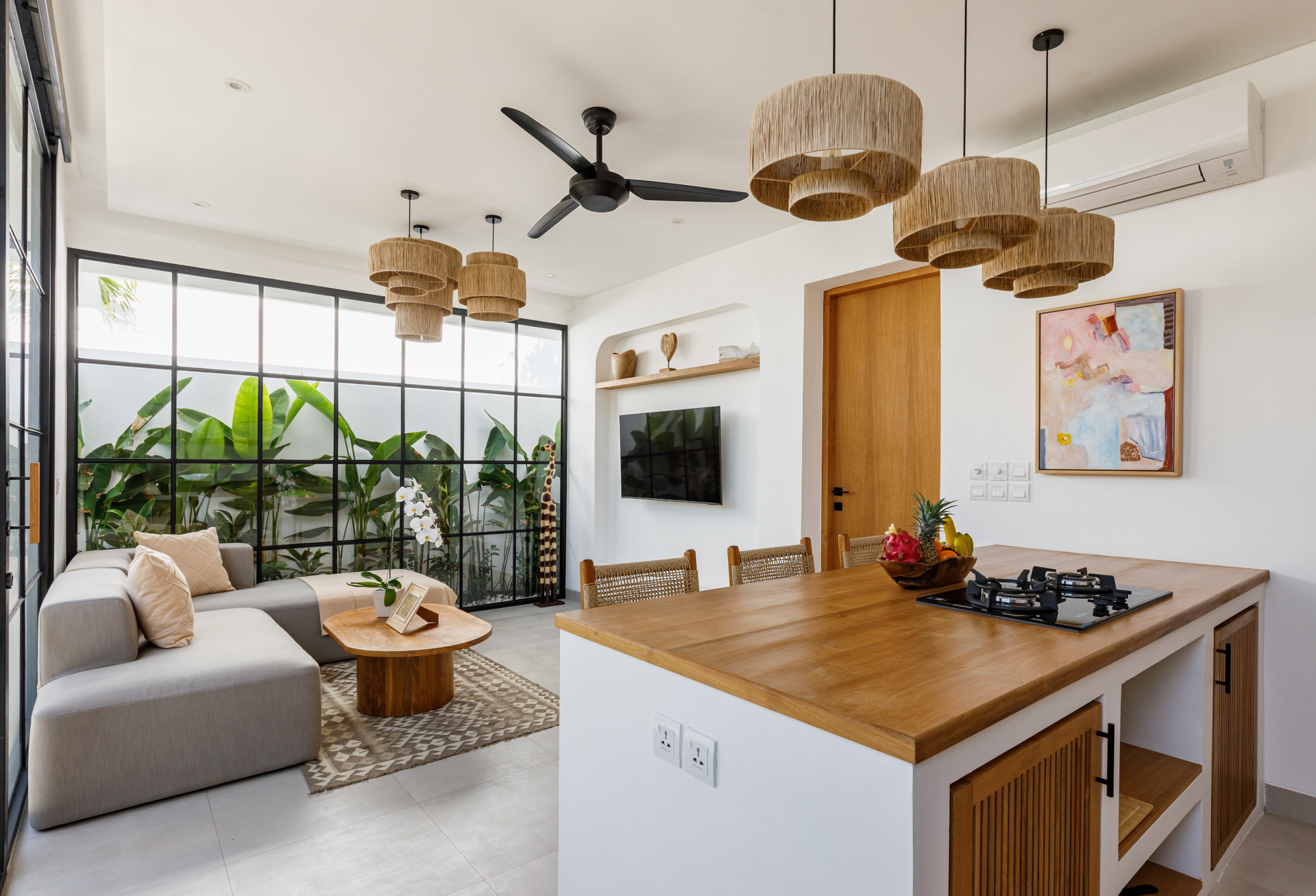 Bright modern living room with gray sofa, wood coffee table, plants, woven pendant lights, and kitchen island