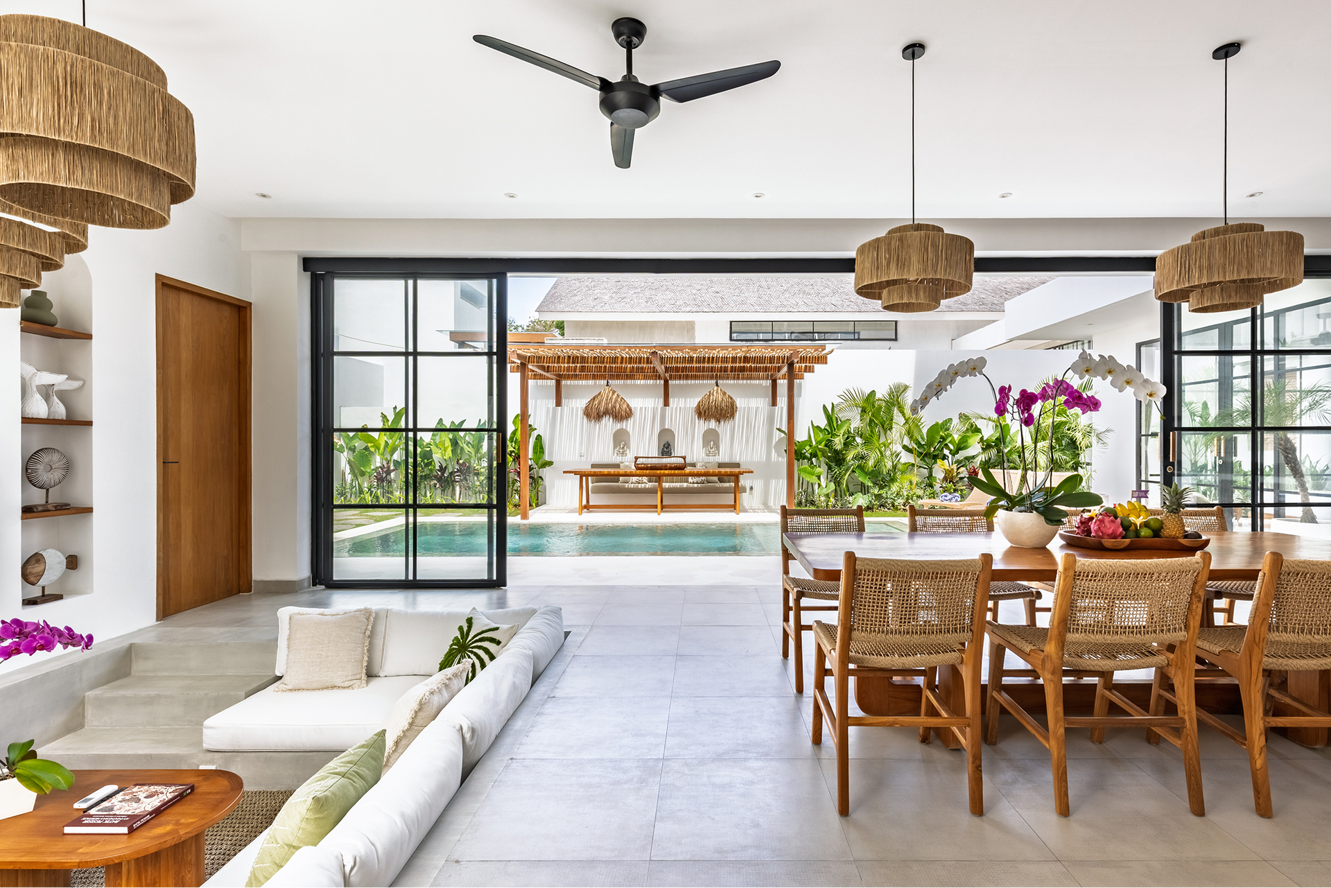 Bright open-air living and dining room overlooking a pool, with wood furniture and tropical decor.