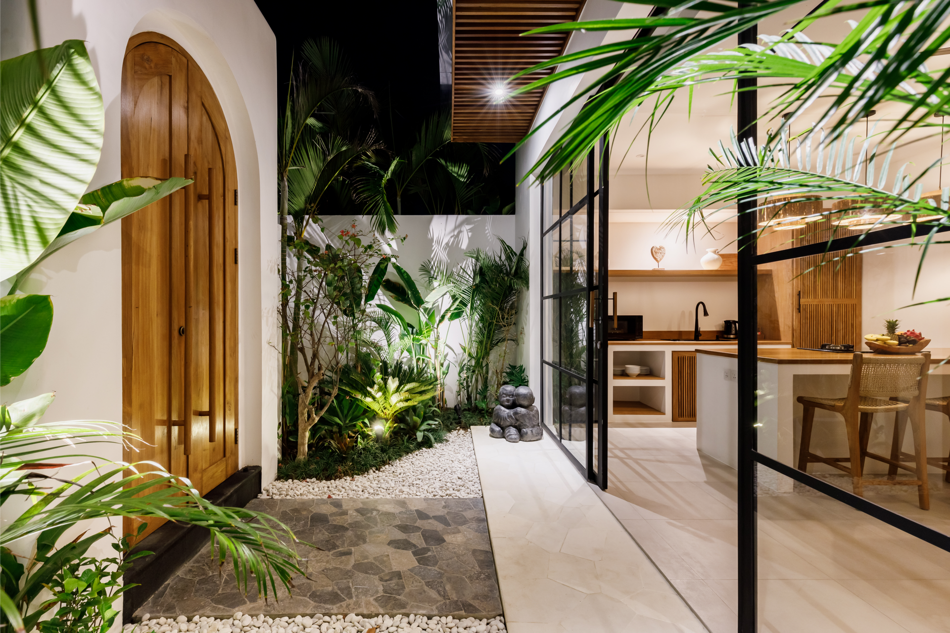 Modern indoor-outdoor hallway with plants, arched wooden door, and warm kitchen seating area