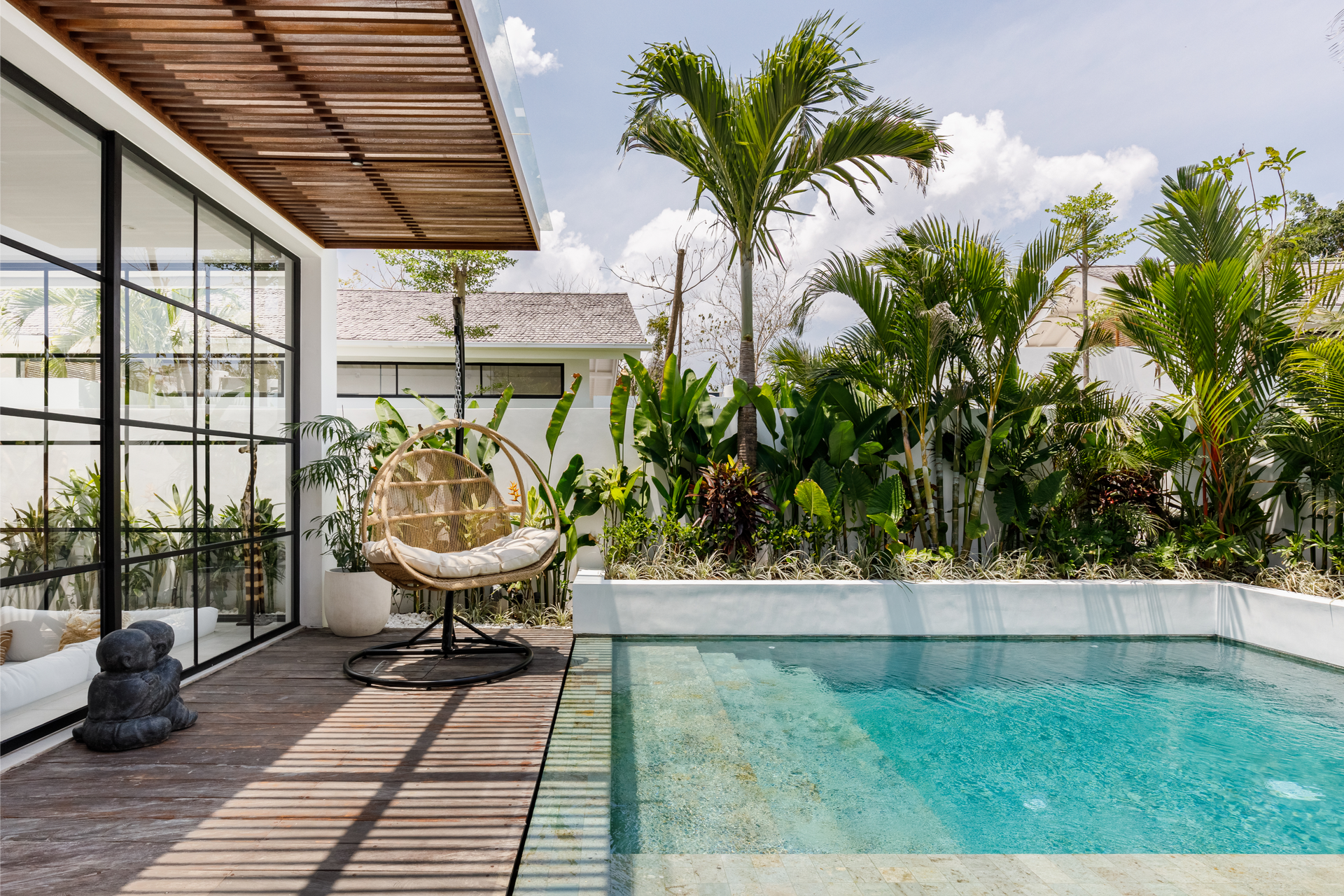 Modern patio with a wicker chair beside a turquoise pool, tropical plants, and a glass-walled house