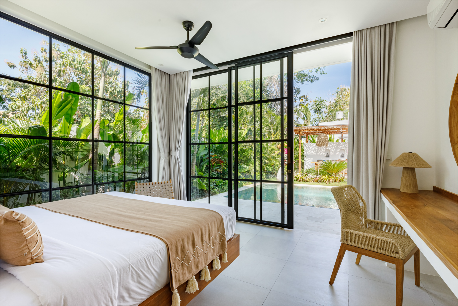Bright bedroom with large black-framed windows, white curtains, a bed, desk, and ceiling fan overlooking greenery.