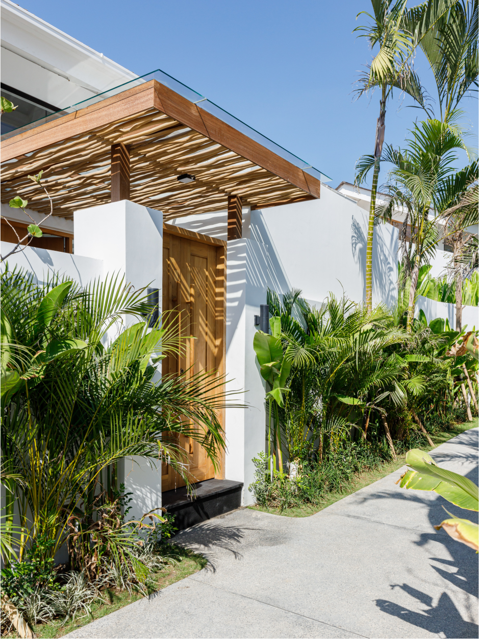 Modern tropical house entrance with wood canopy, white walls, lush greenery, and a paved path