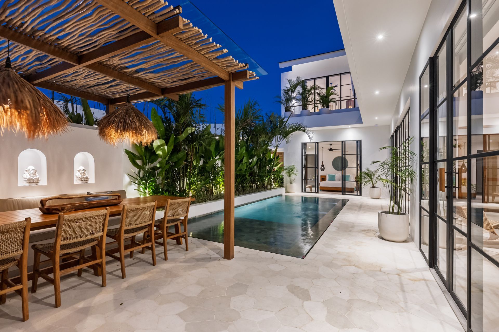 Modern patio with dining table, covered seating, and a narrow pool beside glass-walled rooms at dusk