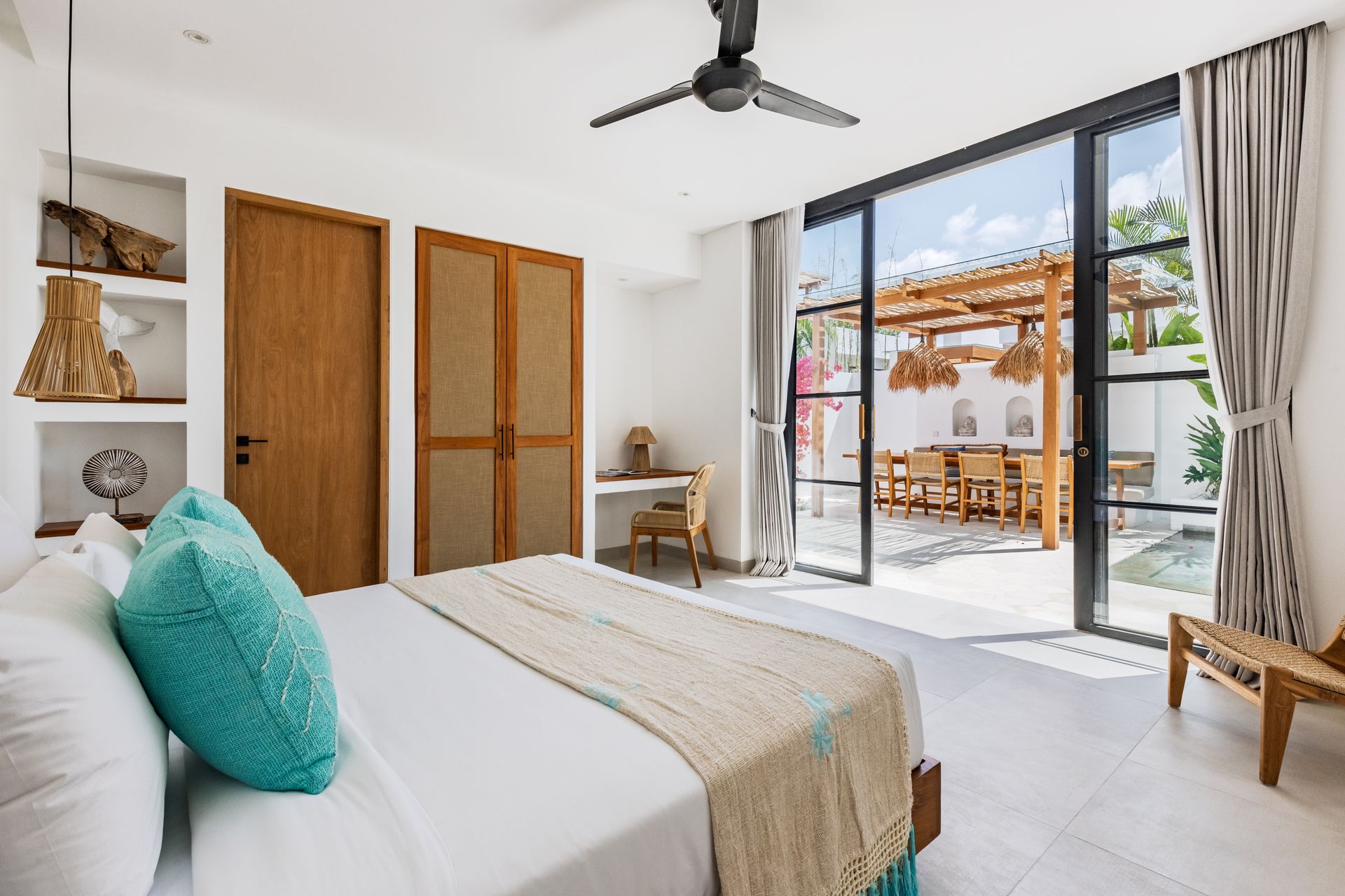 Bright bedroom with white bedding, wooden doors, ceiling fan, and sliding glass doors to a sunny patio.