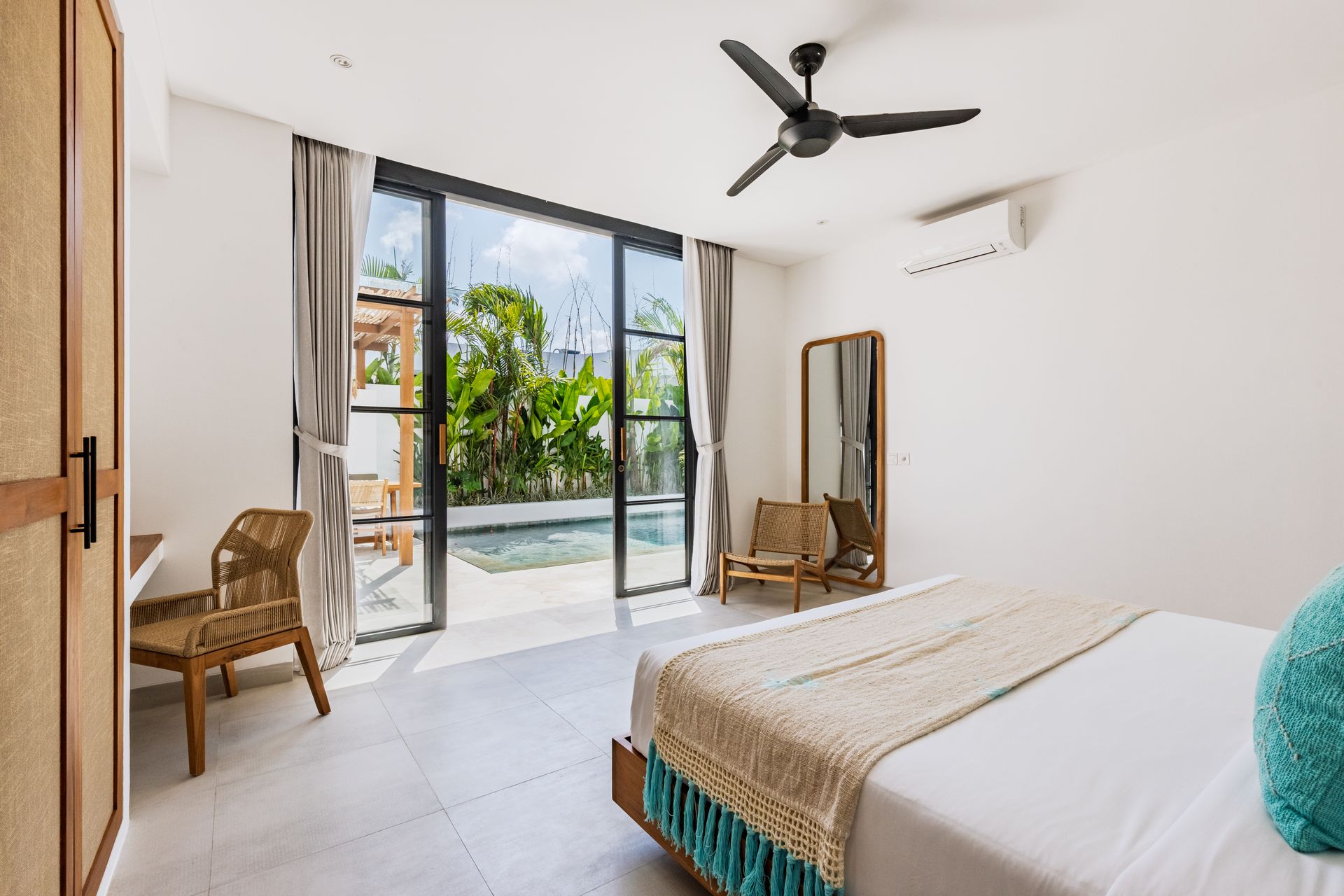 Bright bedroom with sliding glass doors, a bed, wicker chair, full-length mirror, and ceiling fan.