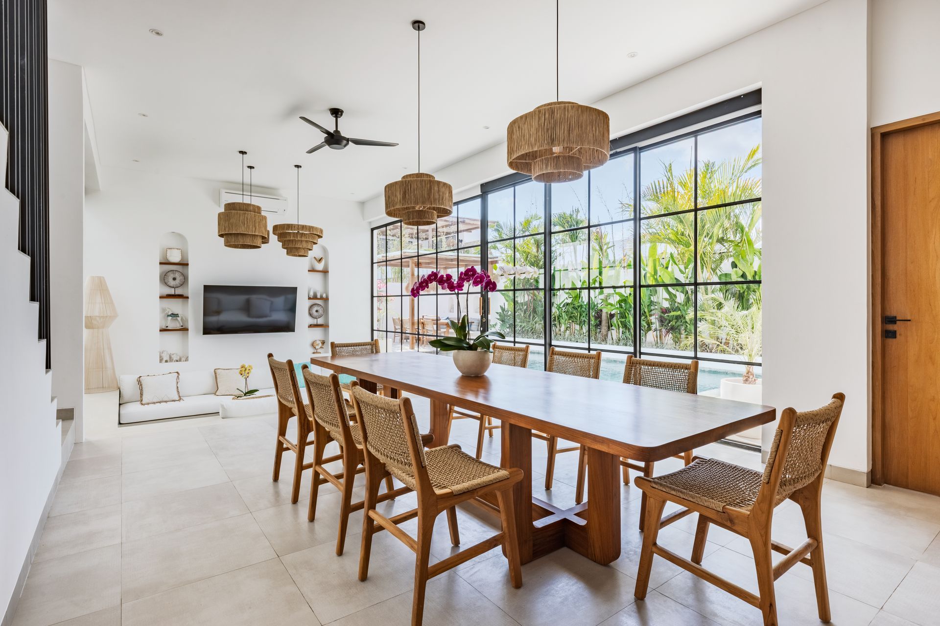 Bright dining room with long wooden table, woven chairs, pendant lights, and large windows overlooking greenery