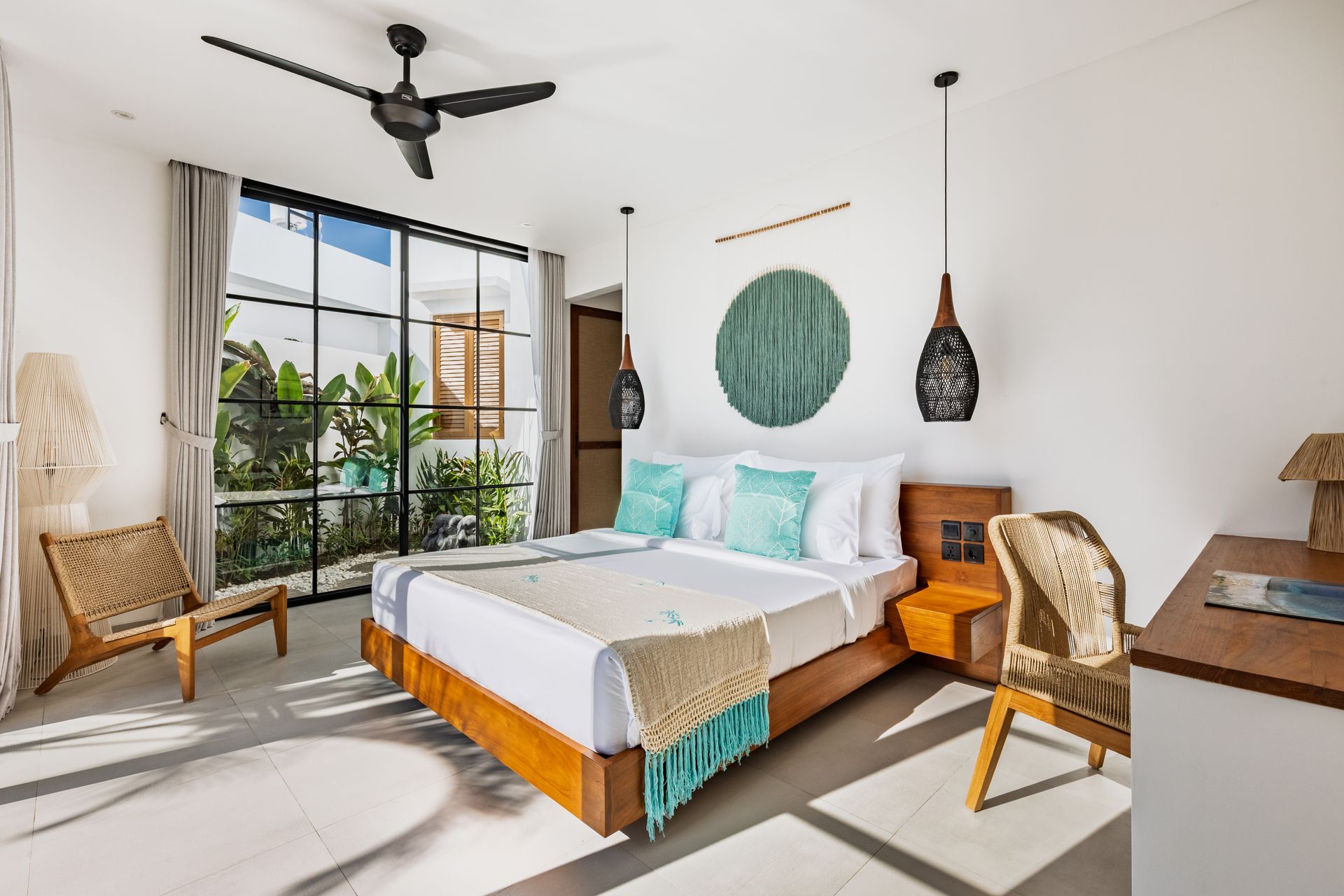 Bright modern bedroom with a bed, wicker chairs, desk, ceiling fan, and large window overlooking greenery