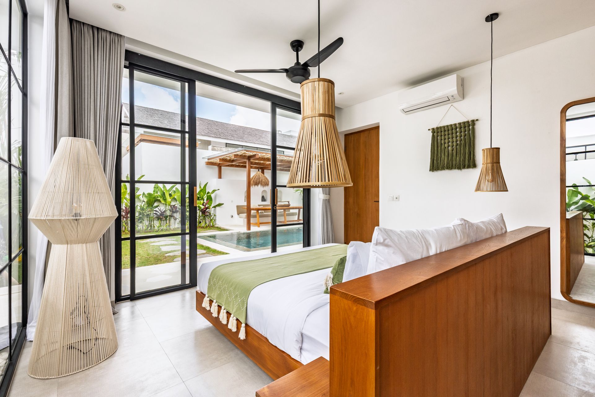 Bright modern bedroom with large glass doors, wooden bed, hanging lamps, and a view of a garden and pool