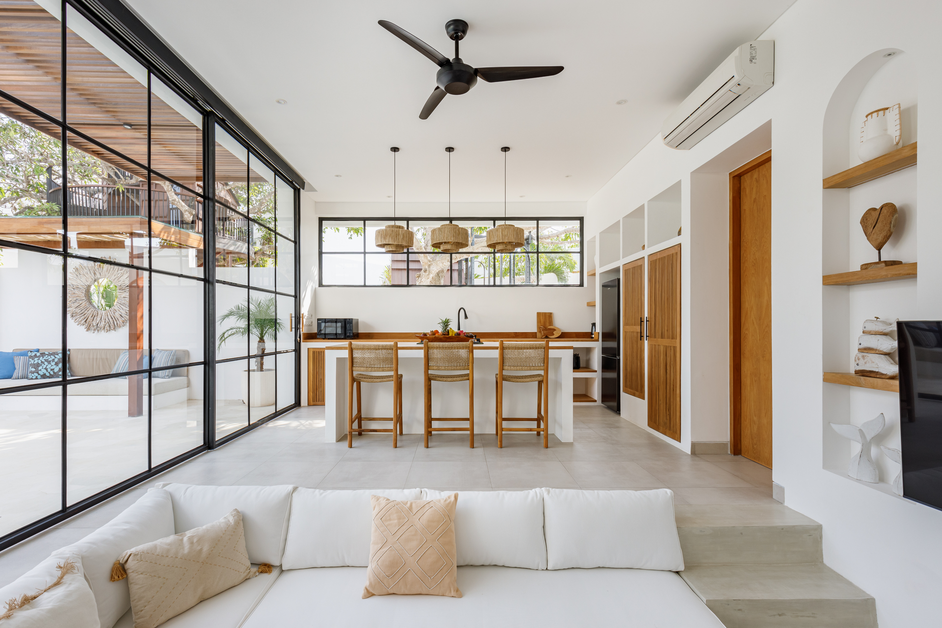 Bright modern open-plan living room and kitchen with white sofa, wooden dining table, and black-framed glass doors.