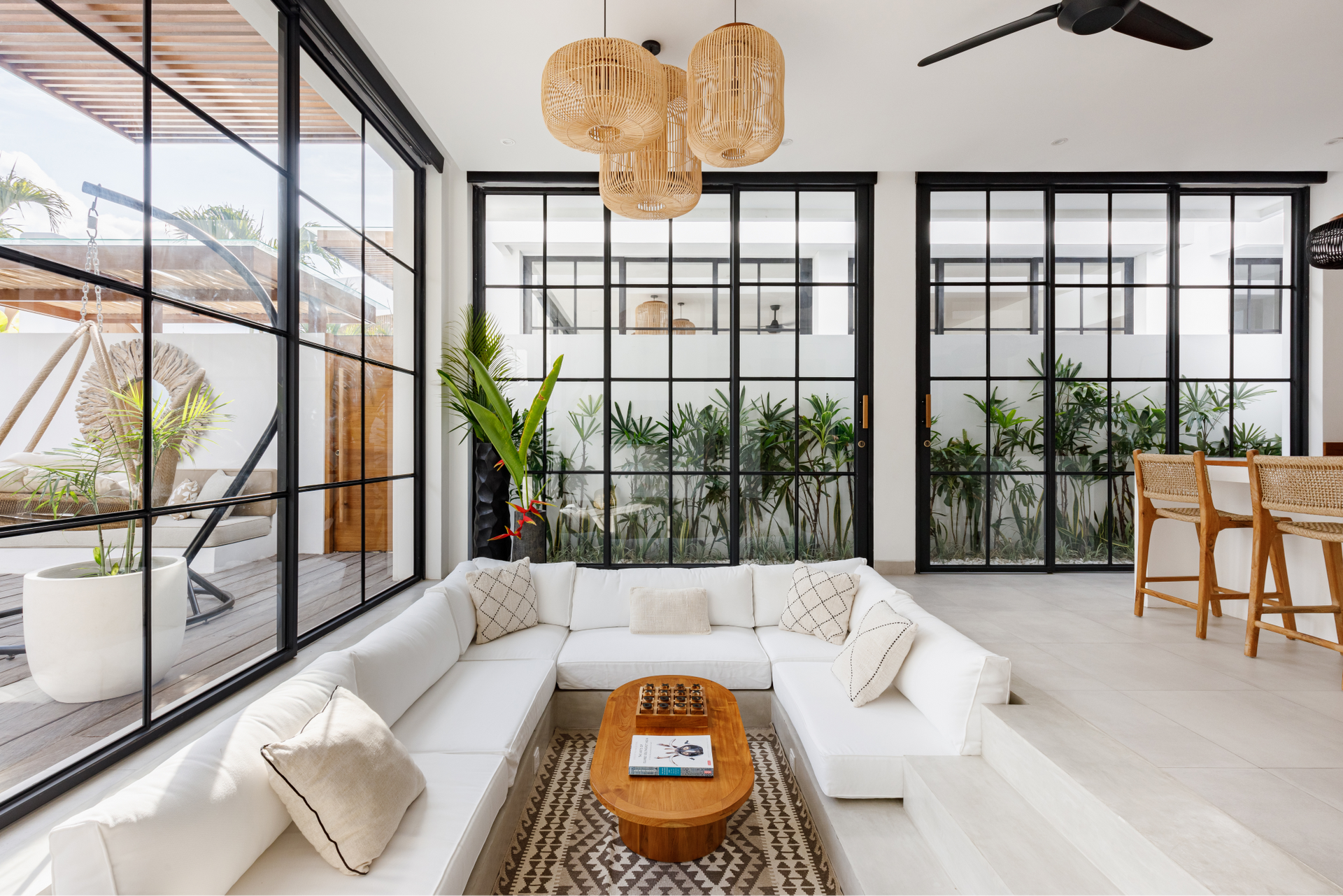 Bright modern living room with white sofa, large black-framed windows, plants, and woven pendant lights