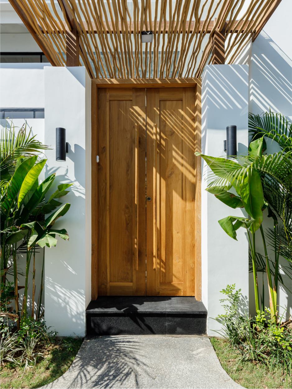 Wooden front door framed by white walls, flanked by lush green plants and a slatted pergola overhead.