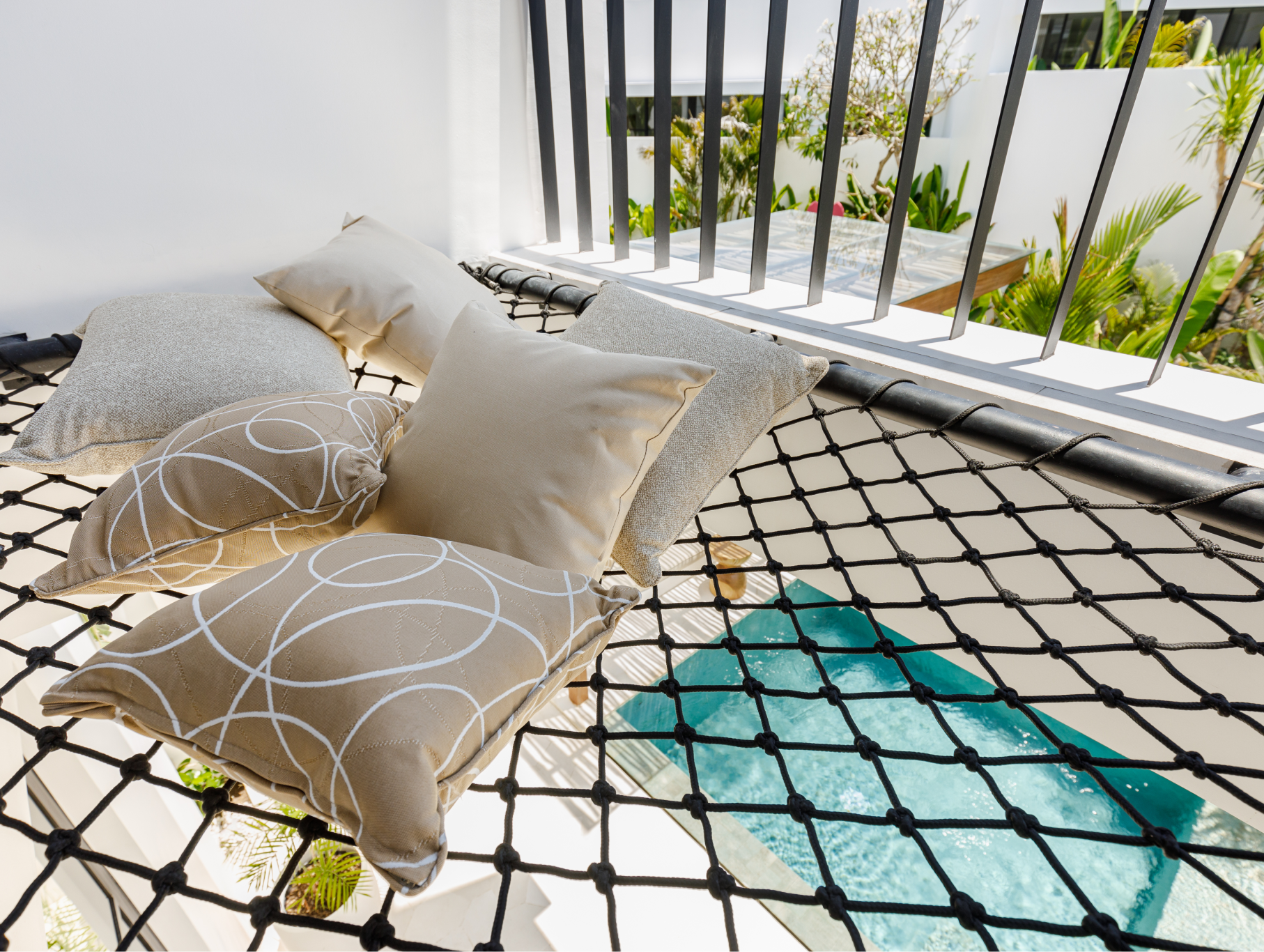 Patterned floor cushions on a netted balcony beside a turquoise rug and railing, with greenery beyond.