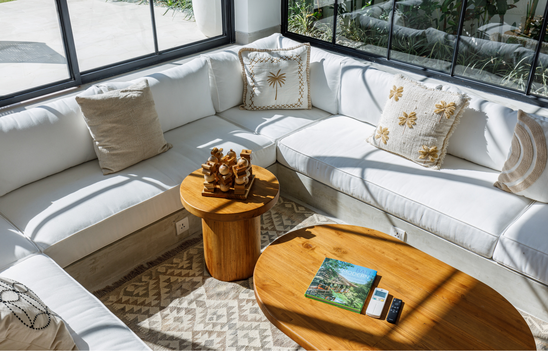 Bright sunroom with a white sectional sofa, wooden coffee tables, and patterned cushions.