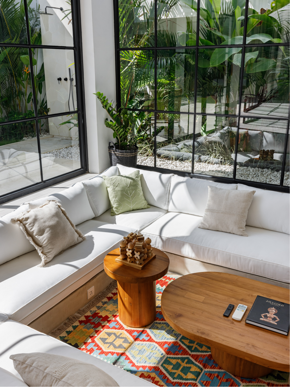 Bright sunroom with white sectional sofa, wooden tables, and colorful rug by large windows