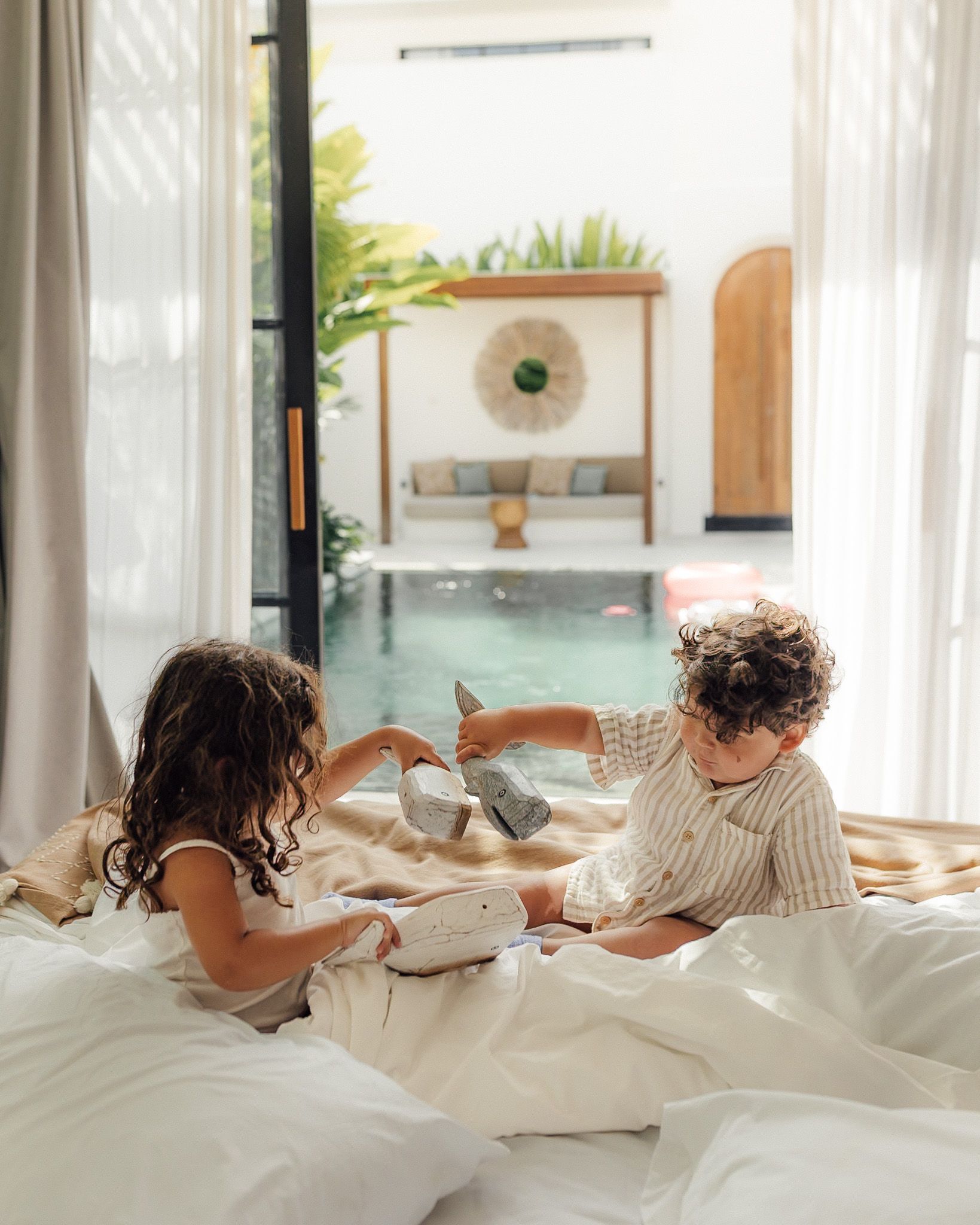 Two children play on a bed beside an indoor pool in a bright, airy room