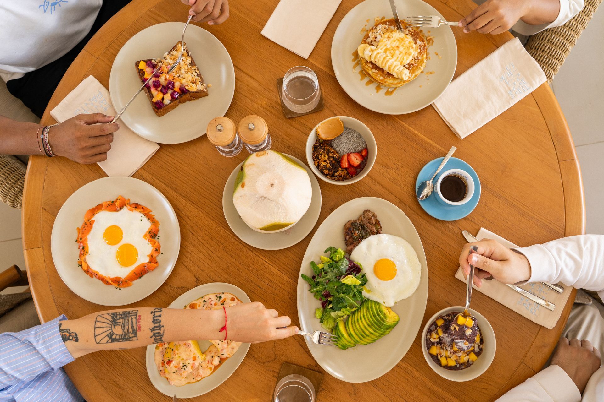 Top-down view of people sharing brunch around a round table with eggs, toast, fruit, and coffee.