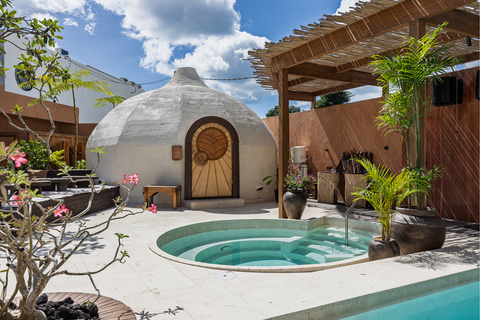 Sunlit courtyard with white dome hut, round hot tub, potted palms, and pergola seating area