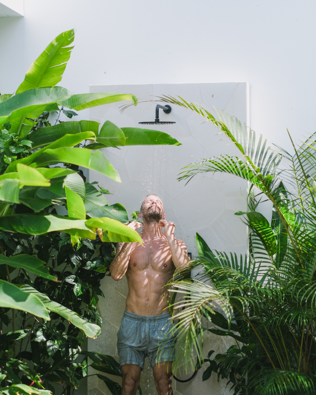 Person standing under an outdoor shower surrounded by lush green tropical plants.