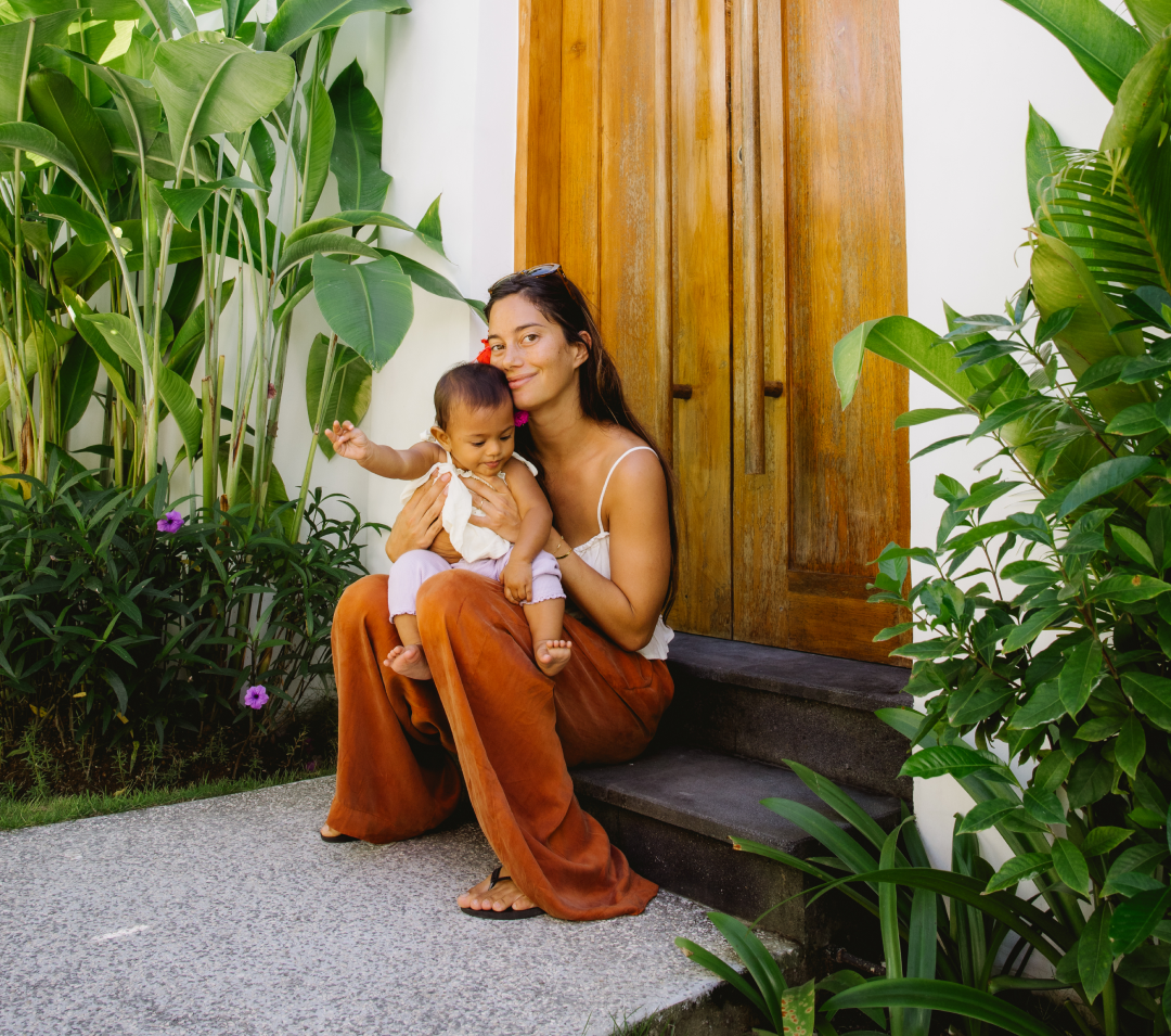 Woman holding a child on outdoor steps beside lush green plants and a wooden door