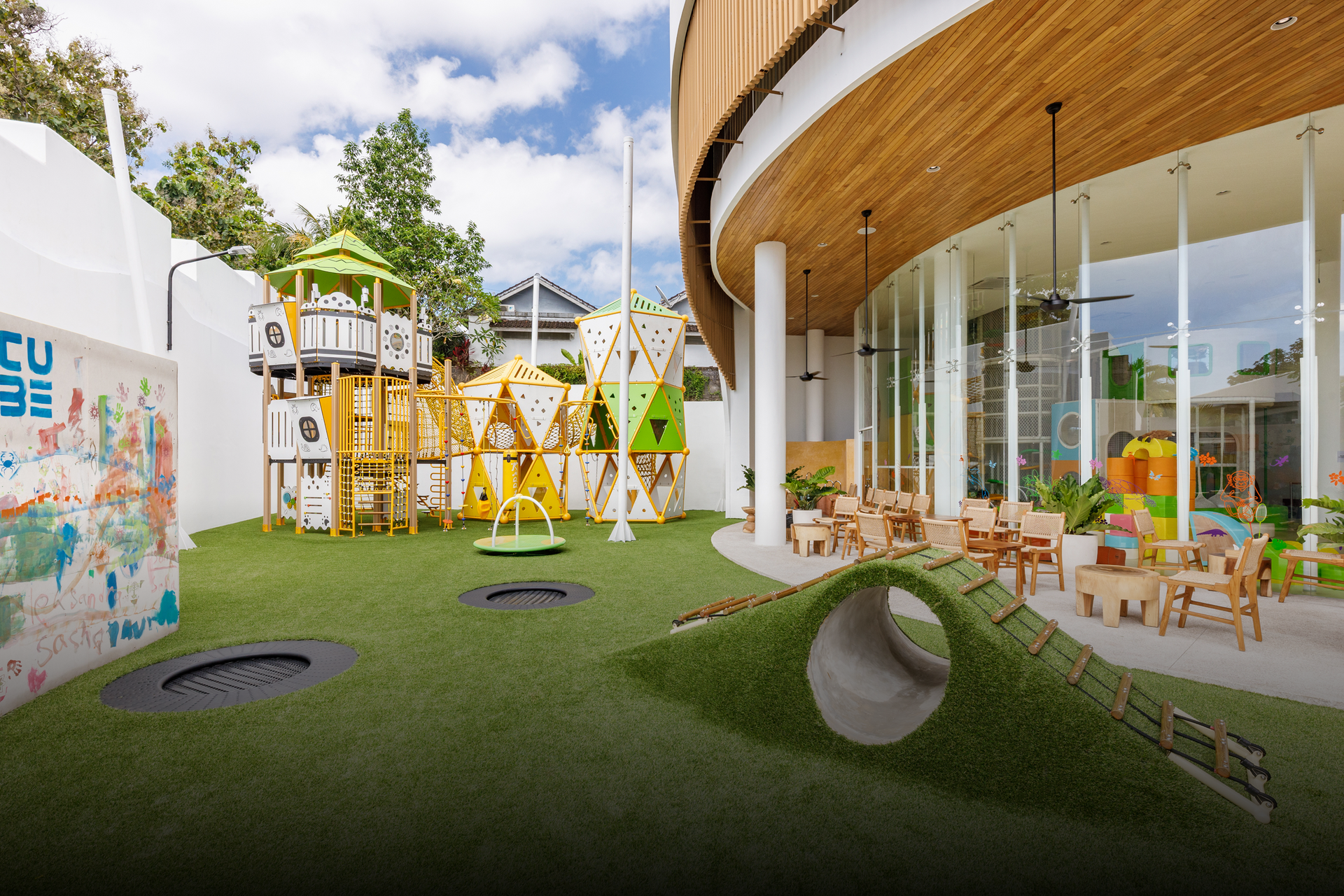 Modern playground with green turf, wooden play structures, and a curved glass building patio nearby