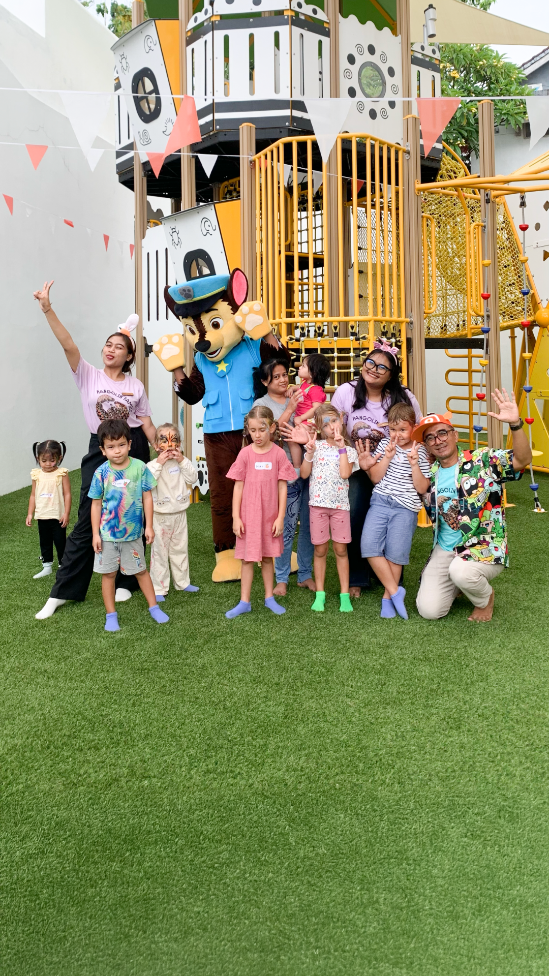 Children posing on a playground lawn beside a yellow play structure and colorful bunting.