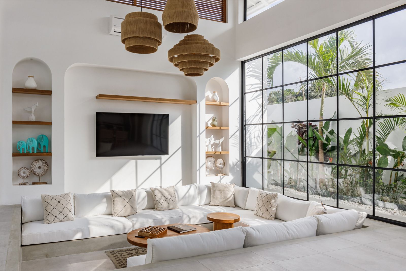 Bright white living room with low seating, woven pendant lights, and a large black grid window overlooking greenery