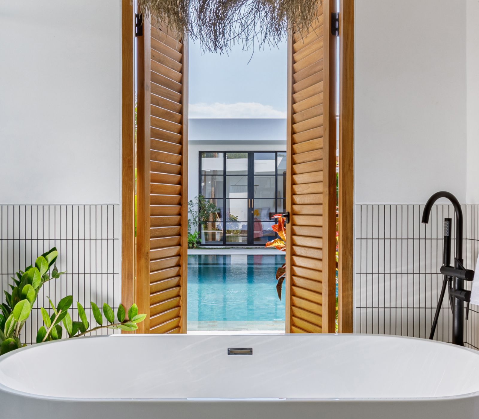 Freestanding tub overlooking a pool through open wooden shutters in a bright modern bathroom
