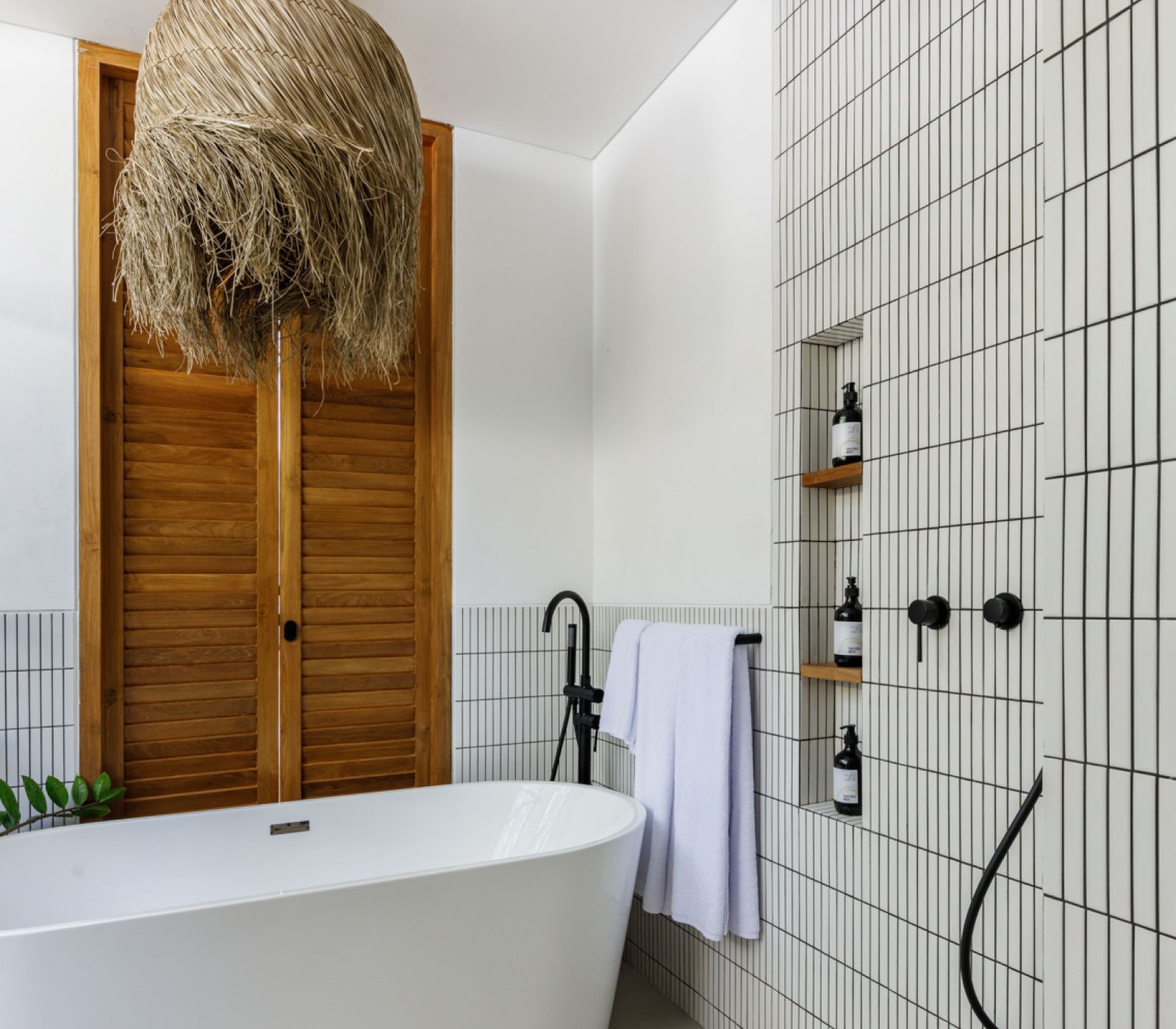 Modern bathroom with freestanding tub, black fixtures, wood shutters, and white grid-tile shower wall