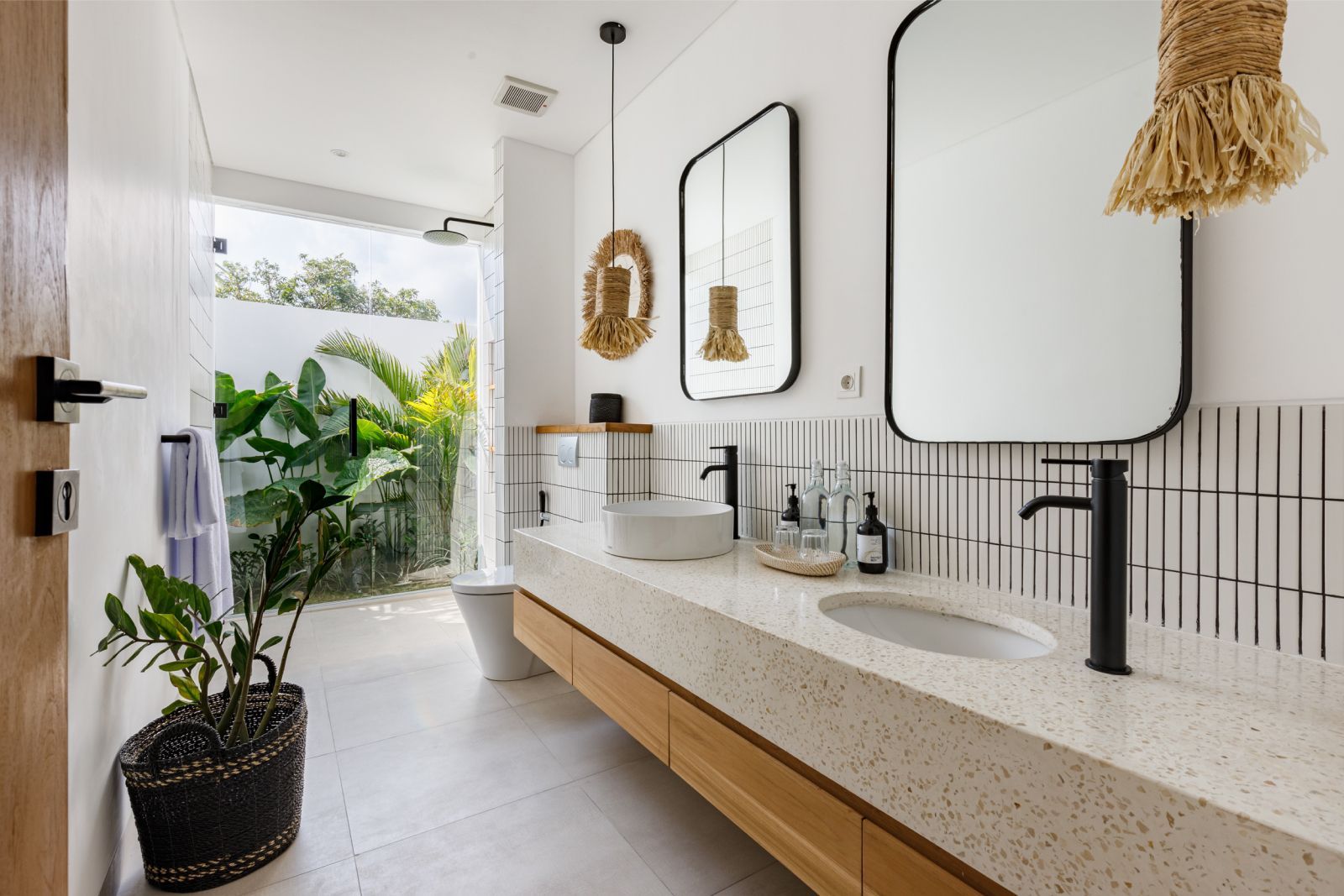 Bright modern bathroom with long wood vanity, speckled countertop, black fixtures, and a view to greenery outside