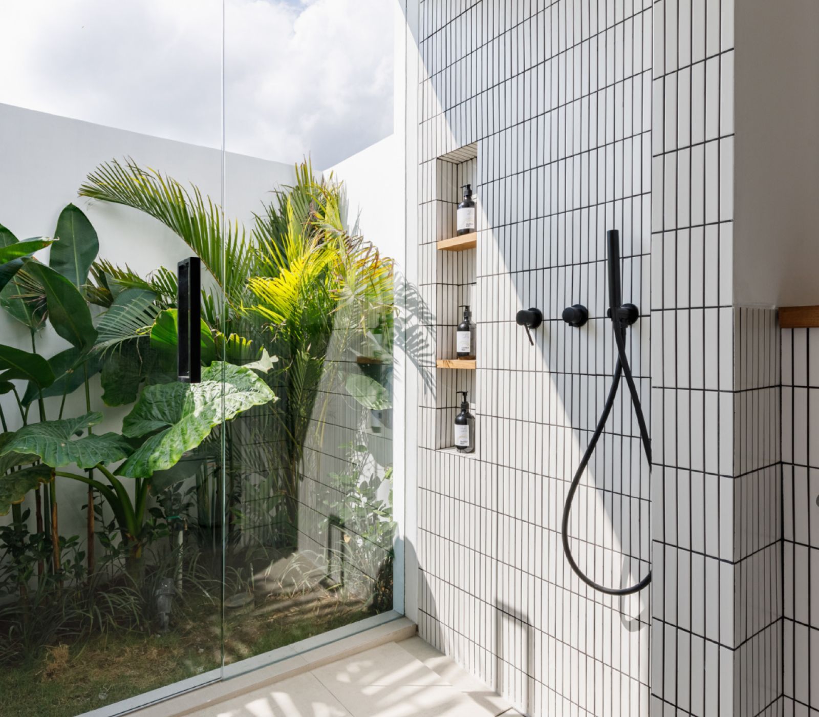Outdoor shower with white tiled wall, black fixtures, and glass partition beside lush tropical plants