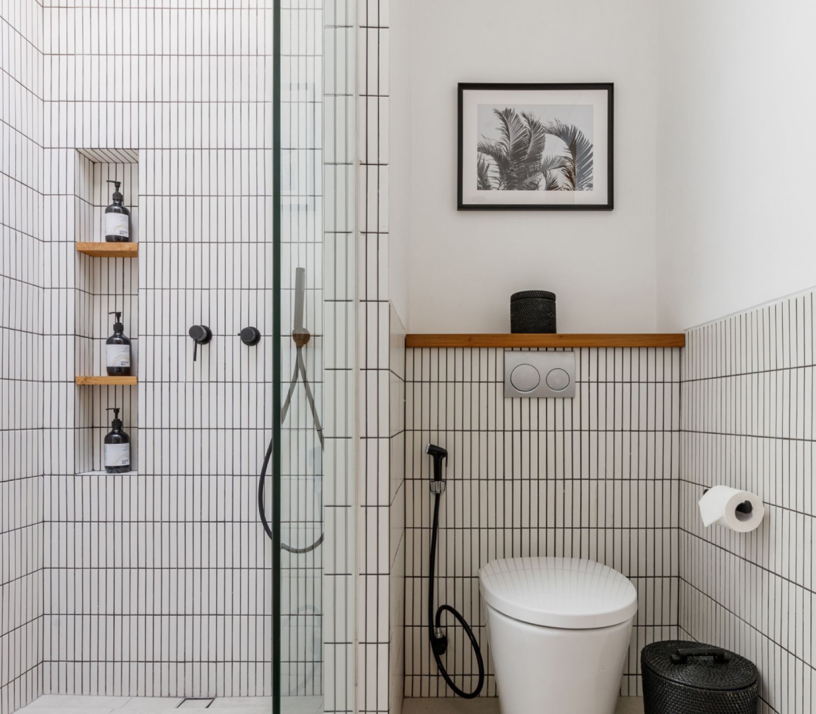 Modern white tiled bathroom with glass shower, toilet, shelves, and framed wall art