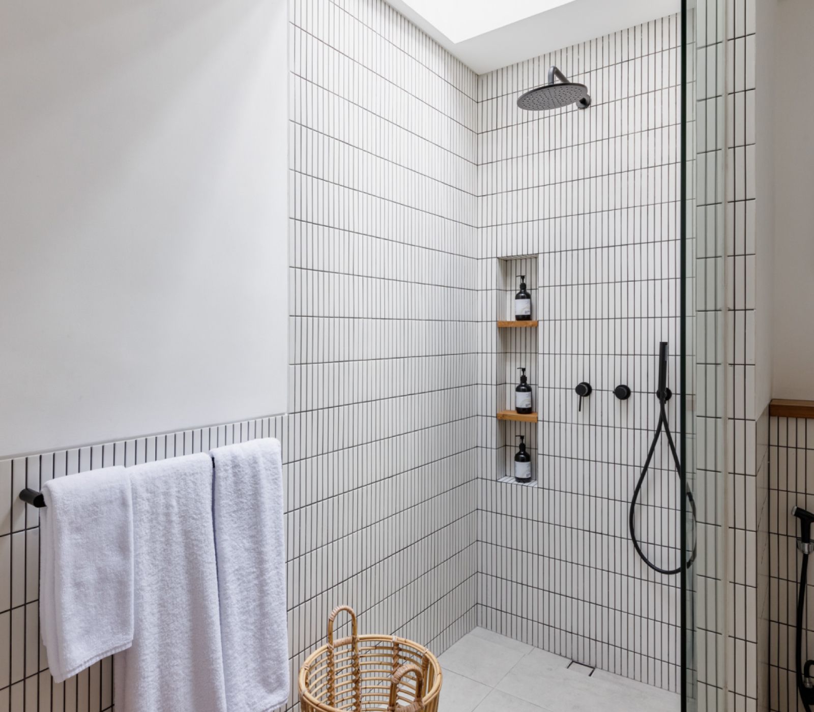 Minimal white-tiled shower with black fixtures, towel rack, shelving, and a wicker basket in a bright bathroom