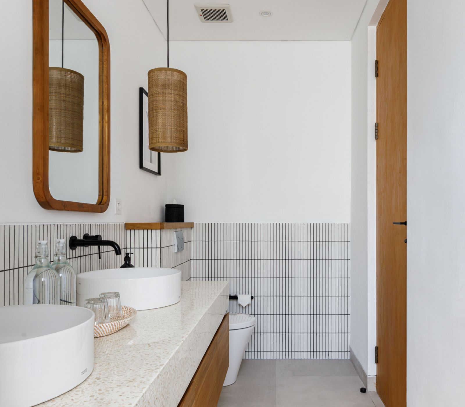 Modern bathroom with double sinks, wood-framed mirror, woven pendant lights, and tiled walls