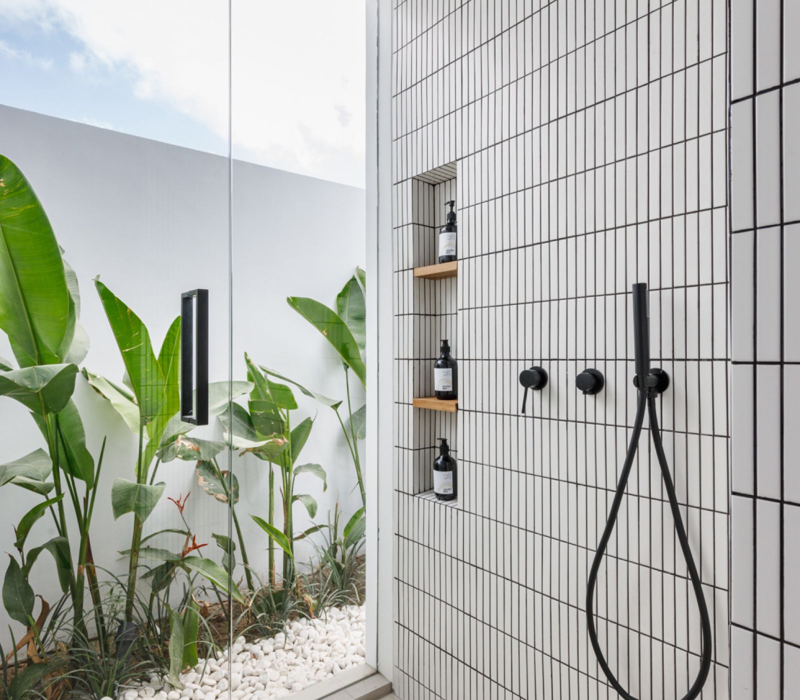 Minimal outdoor shower with black fixtures, tiled wall, mirror, and tropical plants beside white pebbles