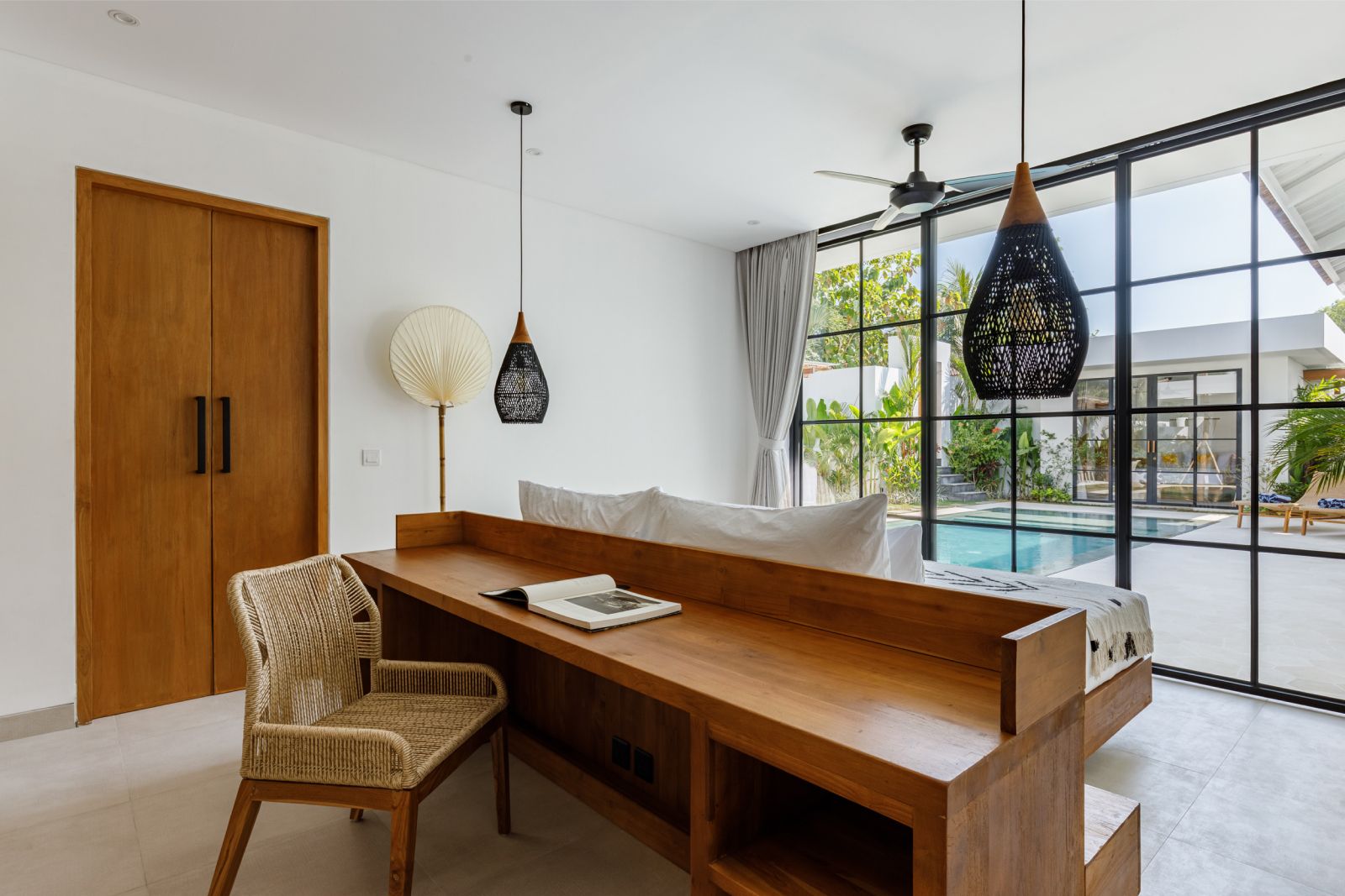 Bright dining room with wooden table, wicker chair, pendant lights, and large windows overlooking greenery