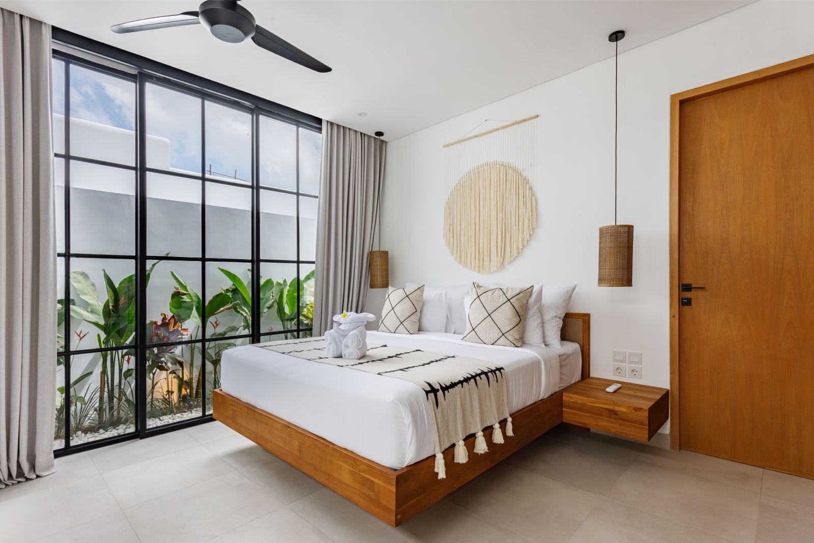 Bright bedroom with a wooden bed, large windows, white decor, and potted plants on a balcony.