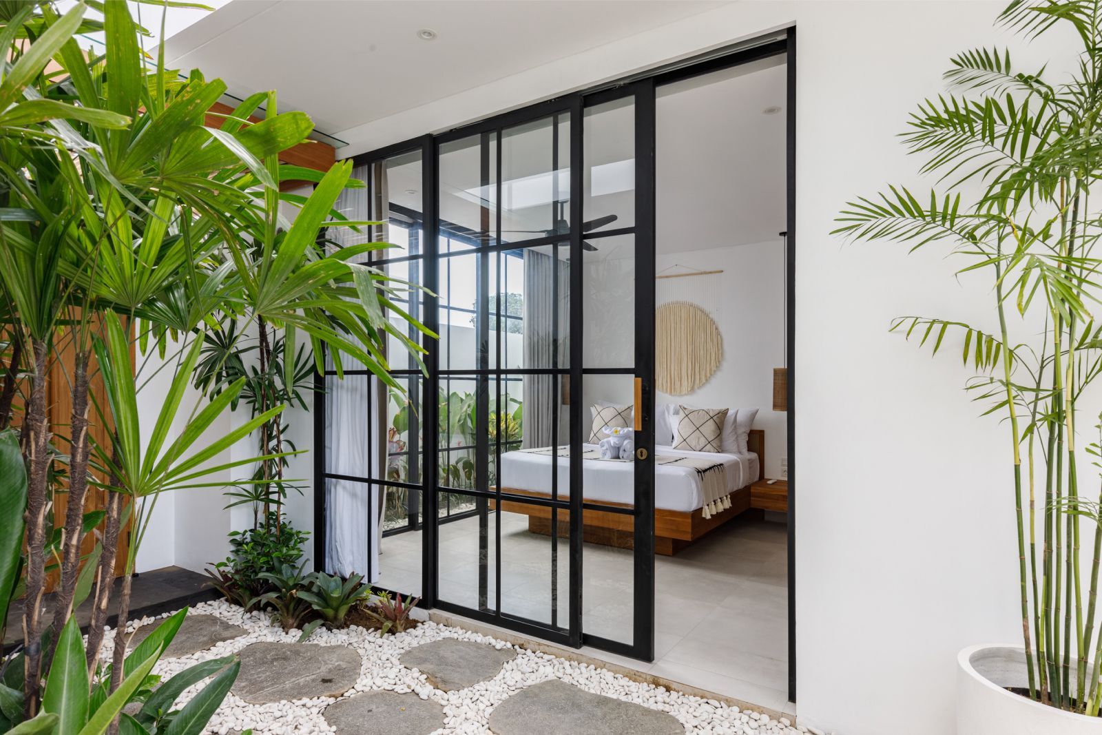 Modern bedroom seen through black-framed glass doors, with white walls and lush indoor plants.
