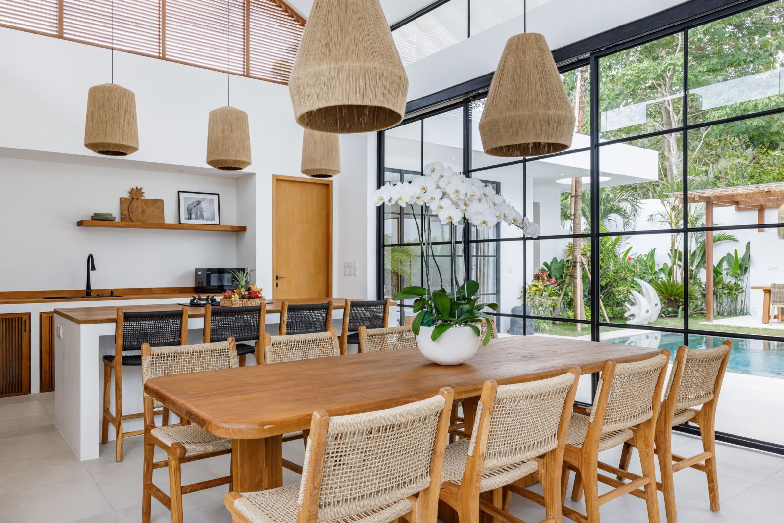 Bright dining room with wicker chairs, wooden table, hanging pendant lights, and large windows to a garden