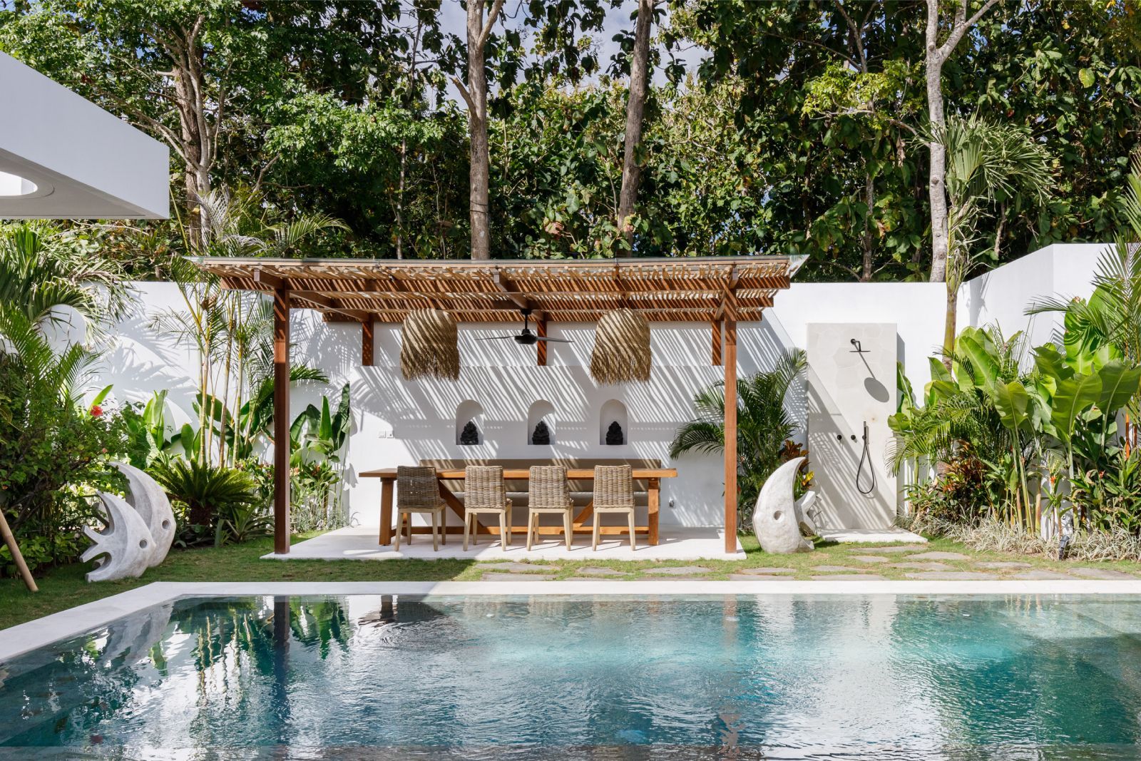 Modern patio dining area beside a turquoise pool, with wooden pergola, table, chairs, and lush greenery