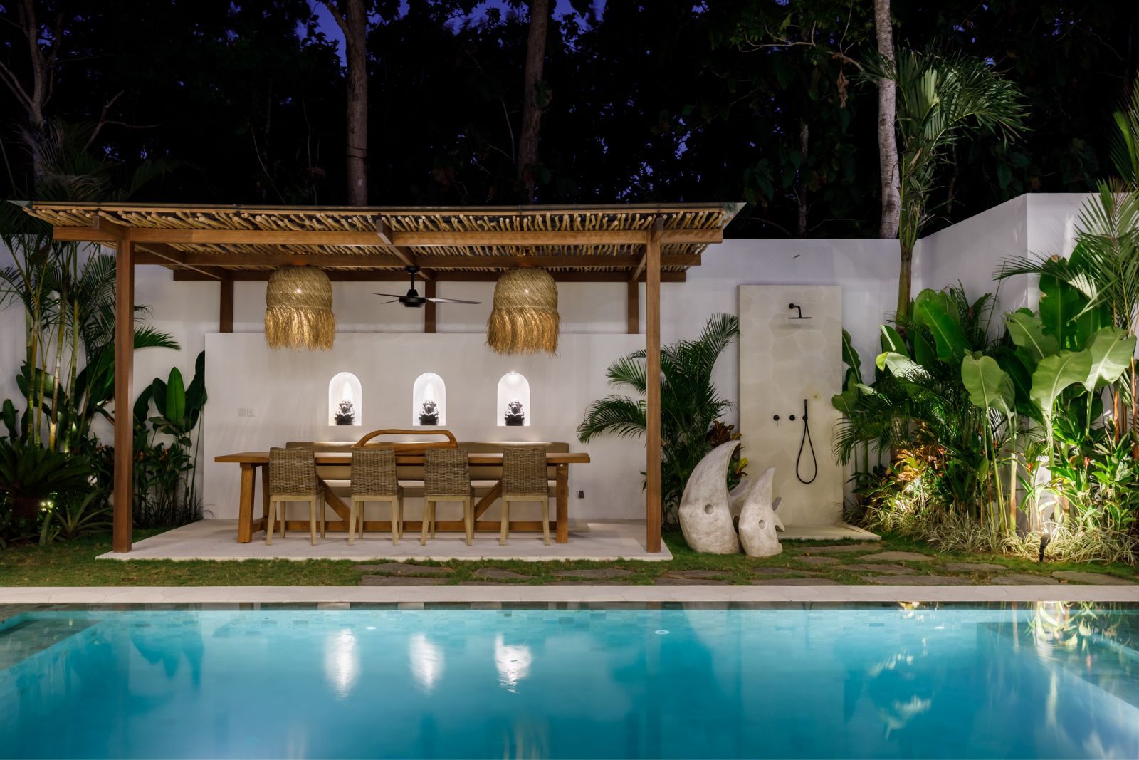 Tropical backyard pool at night with a lit patio dining area, wooden table, and lush greenery