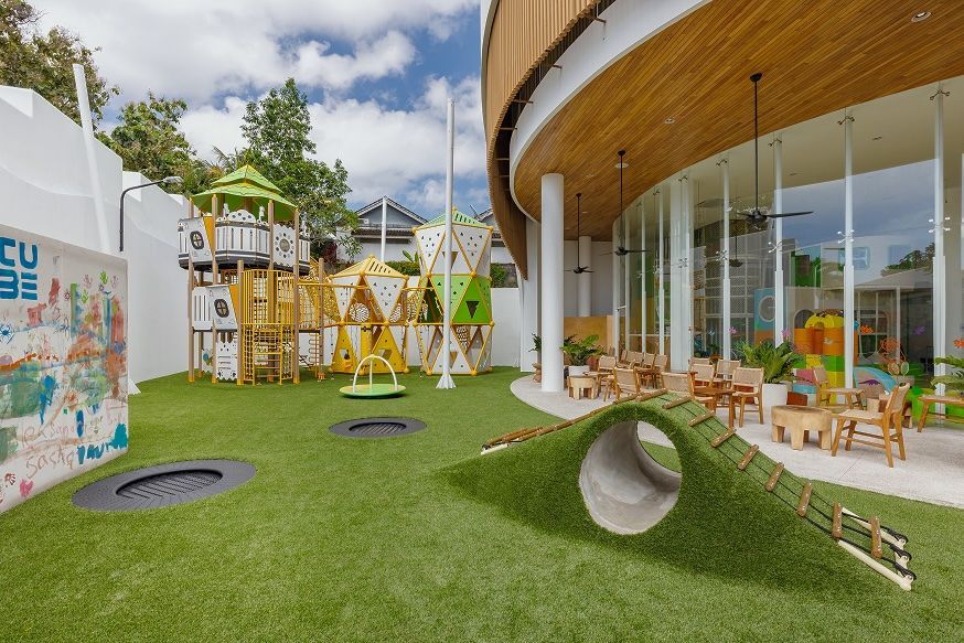 Modern outdoor playground with green turf, wooden play structures, and a curved glass building.