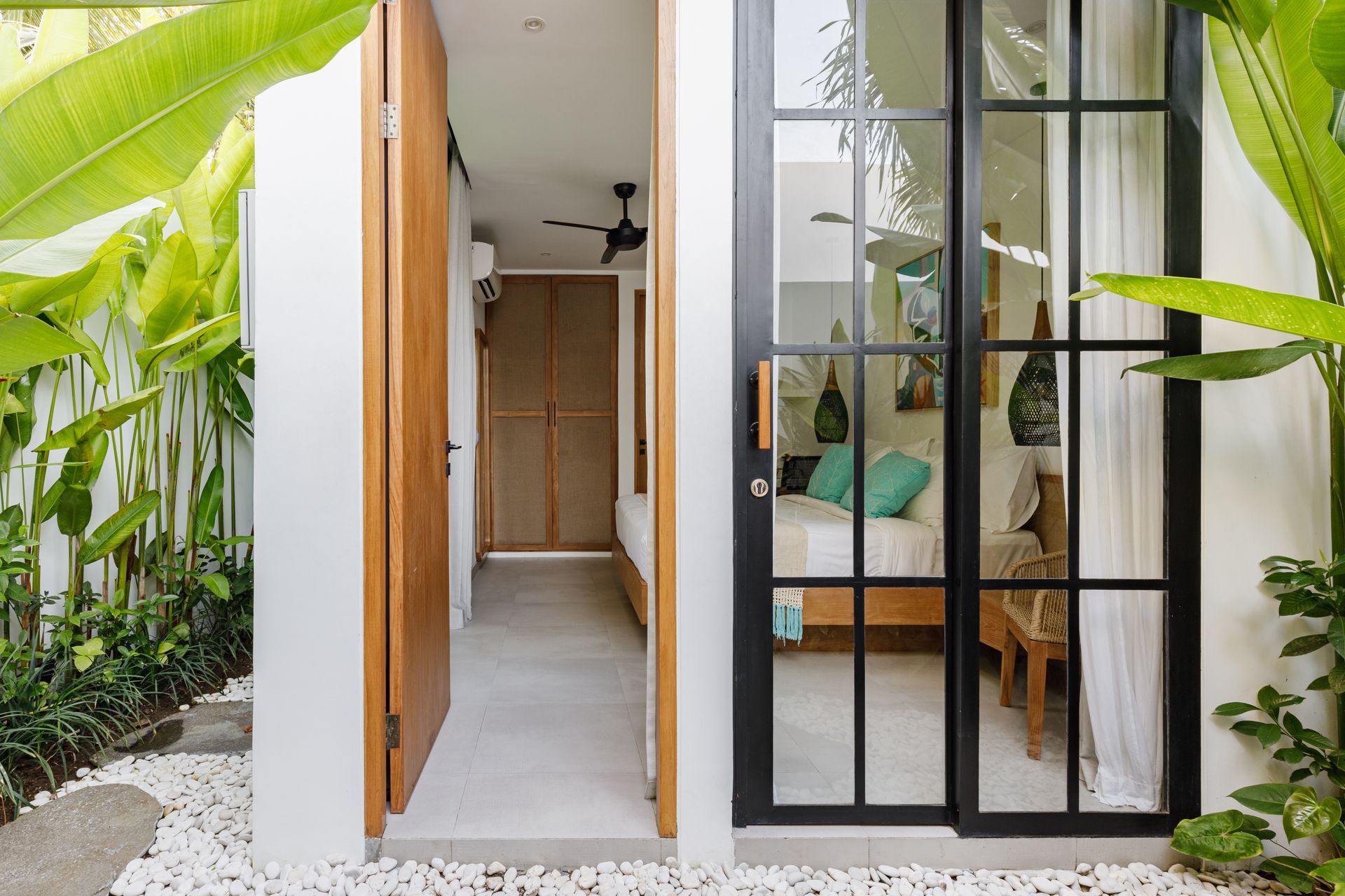 Modern tropical entrance with open wooden door, black-framed glass wall, and lush greenery outside